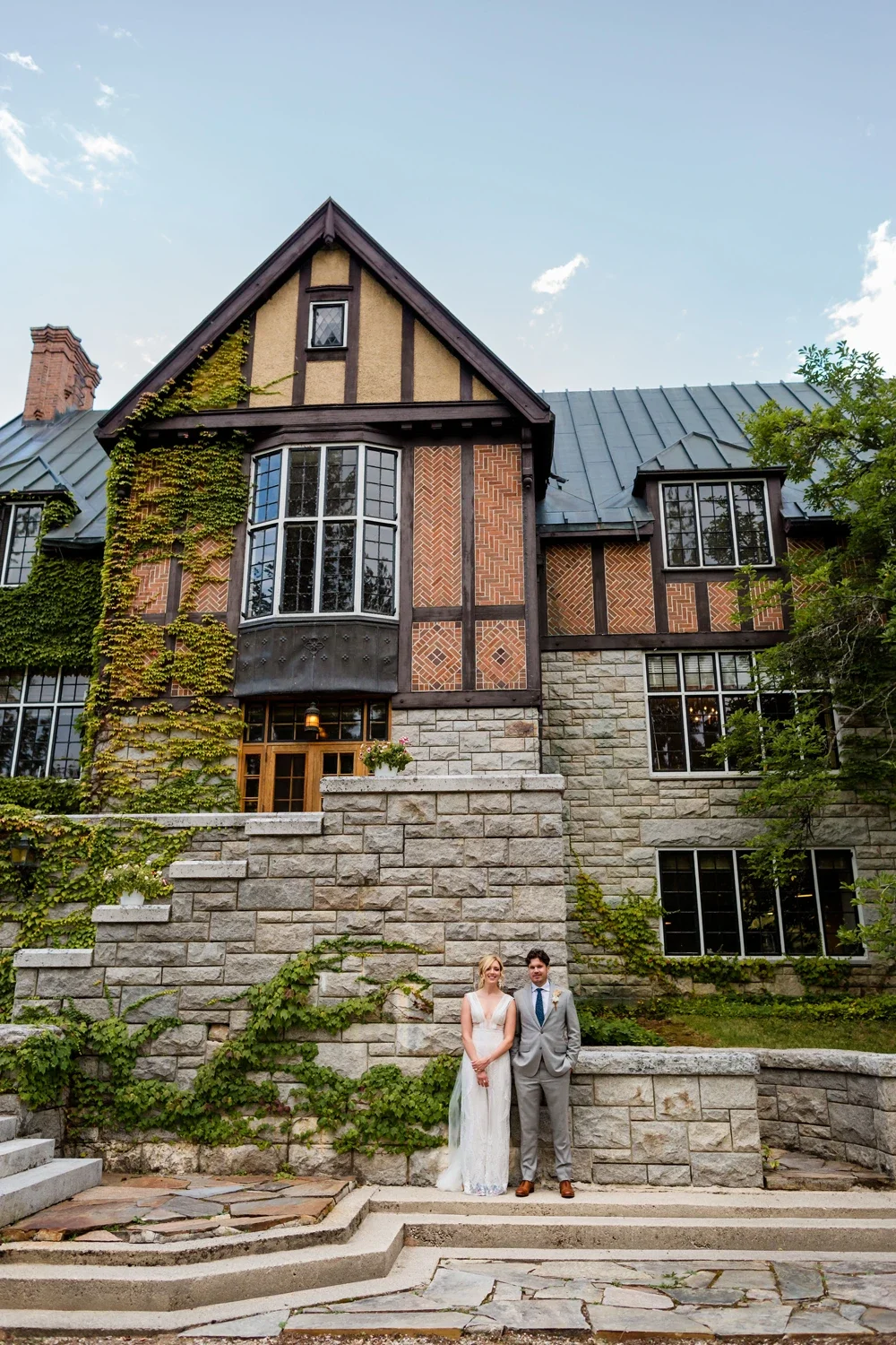 A bride and groom standing outside a large, historic house with stone steps and brick and timber exterior, surrounded by greenery.