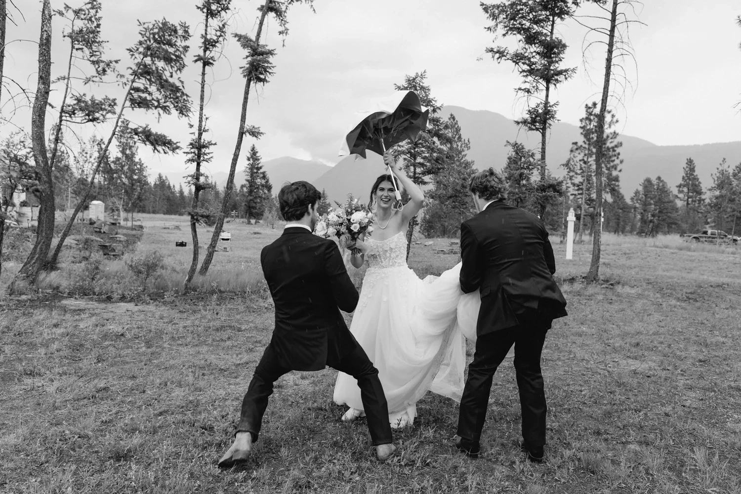A bride holding a bouquet and an umbrella is smiling and posing with two men in suits outdoors in a grassy, wooded area with mountains in the background.