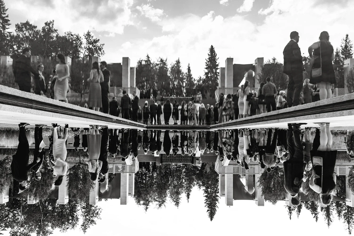 Black and white photo of a large wedding or formal gathering outdoors, with many people dressed in formal attire, some talking in groups. The image has a reflection effect making it appear upside down, with the ground and the sky mirrored.