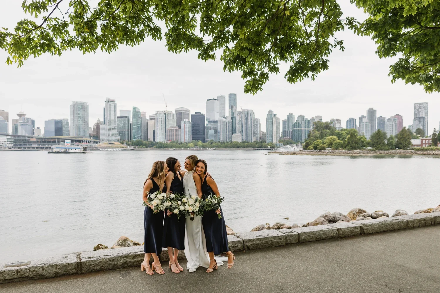 A bride and three bridesmaids standing by a river with a city skyline in the background, holding bouquets of white flowers.