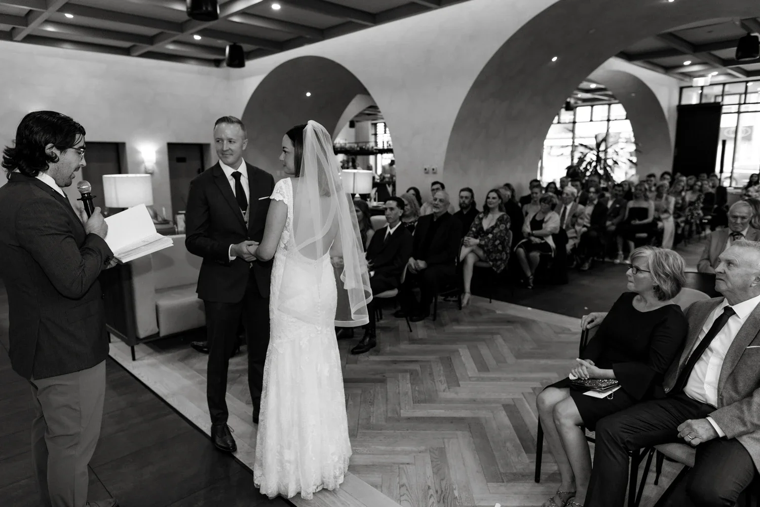 A black-and-white photo of a wedding ceremony with a officiant reading from a book, a couple holding hands in wedding attire, and seated guests watching attentively.