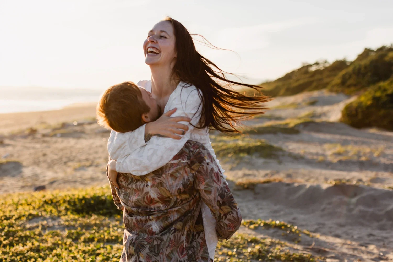 A woman hugging a young boy on a beach at sunset, both smiling and embracing each other.