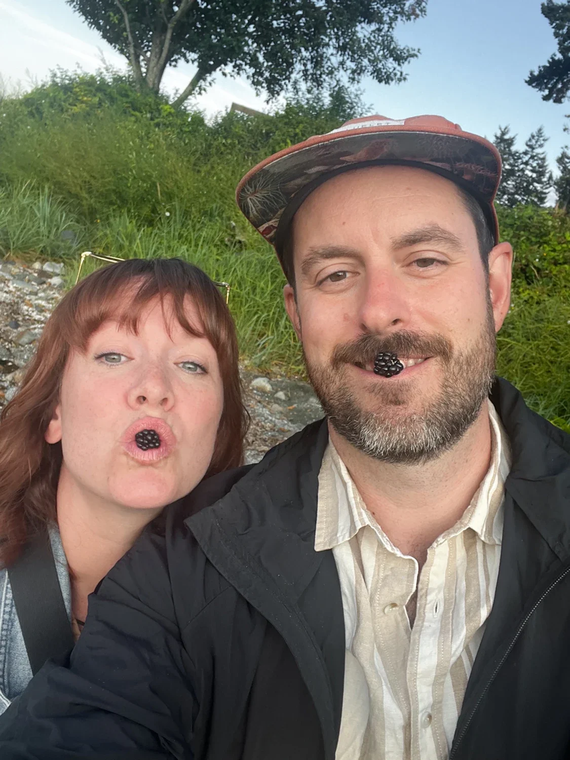 A man and woman taking a selfie outdoors with blackberry in their mouths, wearing casual clothing and surrounded by greenery.