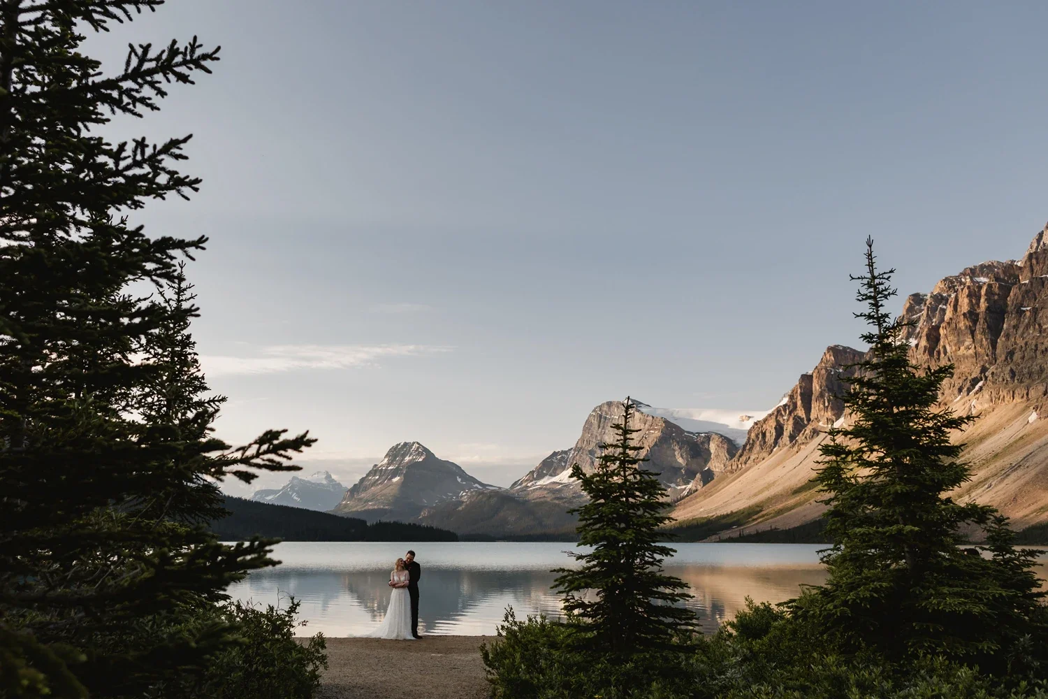 A couple in wedding attire standing on the shore of a lake, surrounded by trees with a mountain range in the background, during daylight.