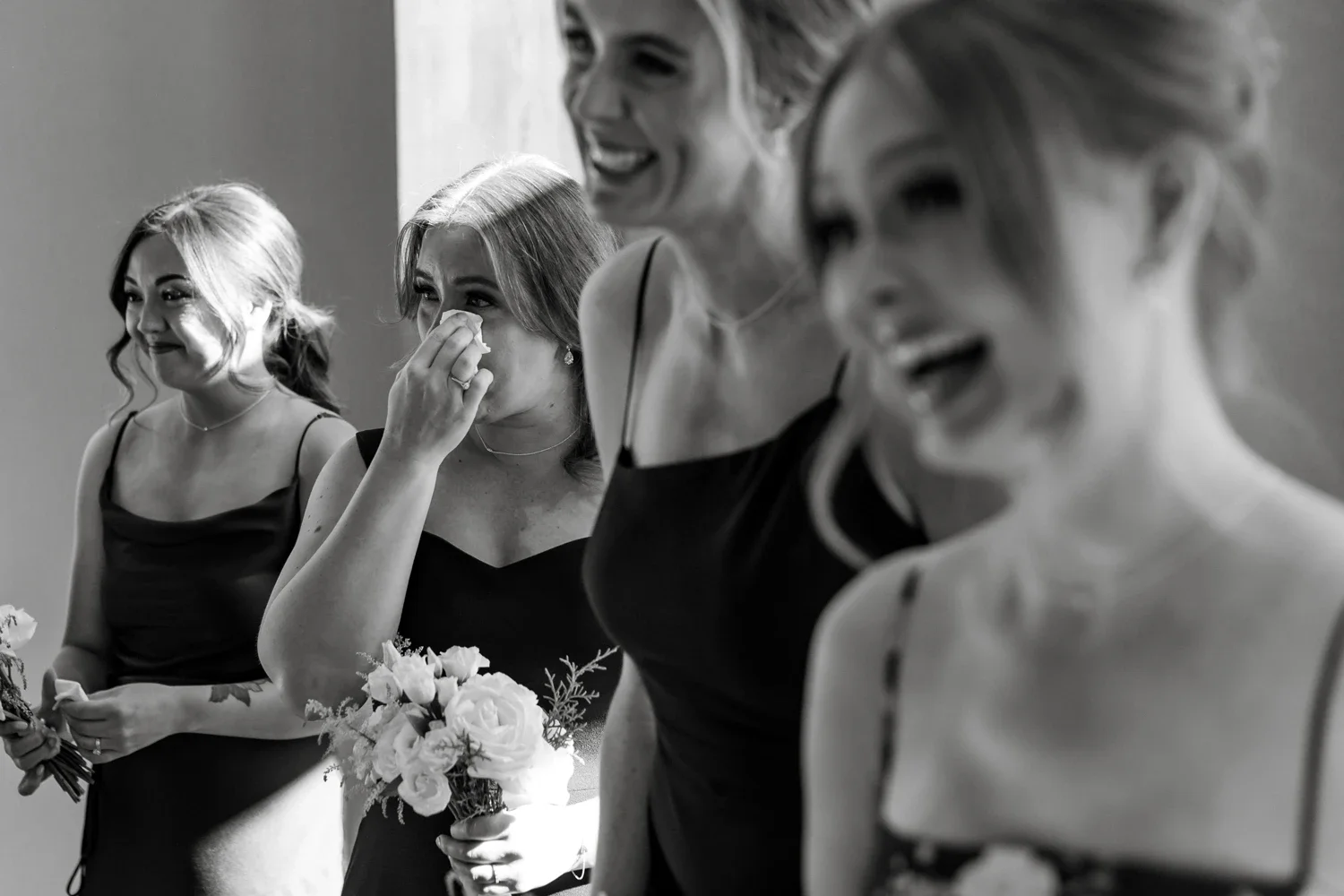 Four women dressed in black dresses, two holding bouquets, are seen laughing and smiling during a heartfelt moment, with one woman wiping tears.
