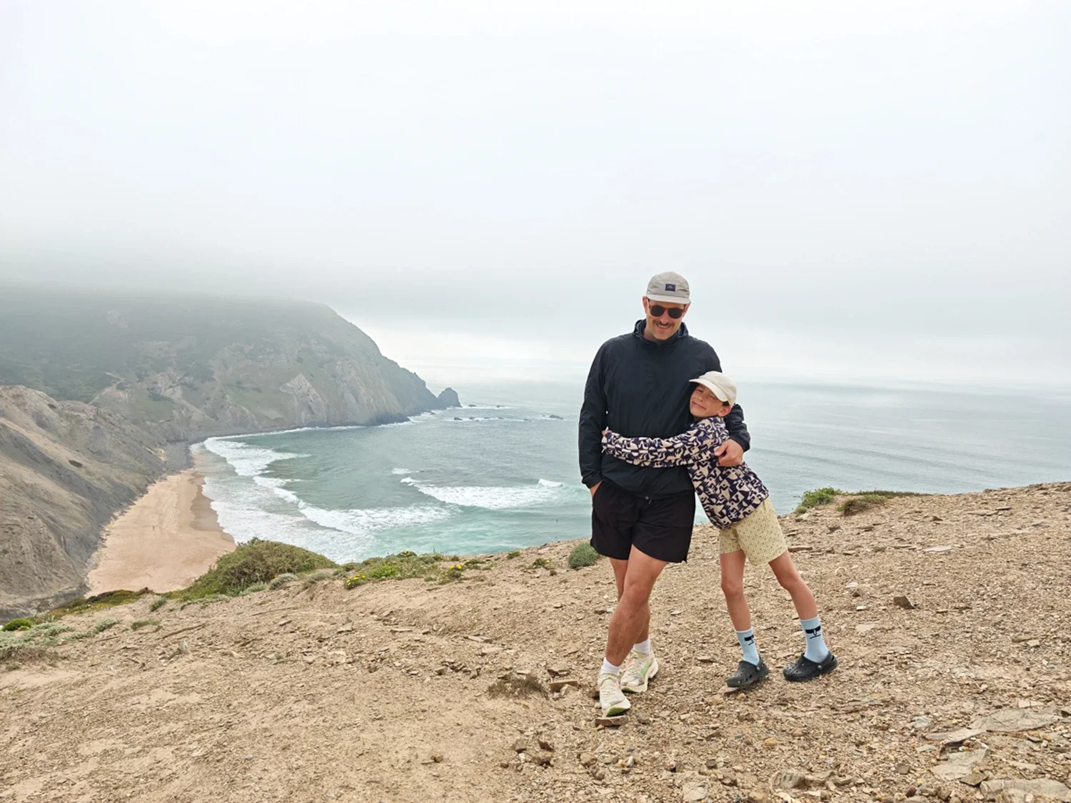 A man and a young girl embracing on a cliff overlooking a beach and ocean with overcast skies.