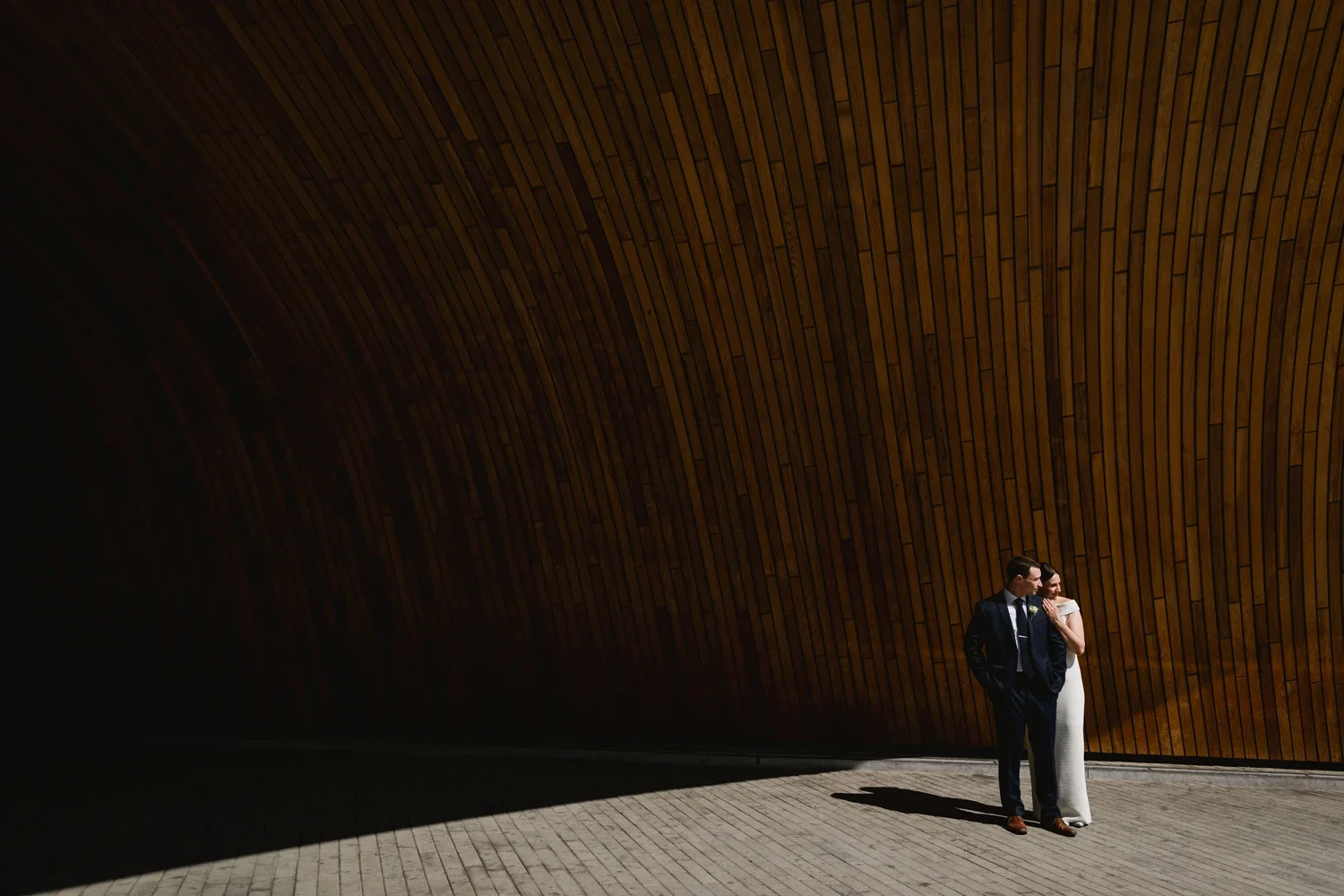 A couple in wedding attire standing close together against a large, curved wooden wall. The groom is in a dark suit and the bride in a white gown, with both casting shadows on the light-colored brick ground.