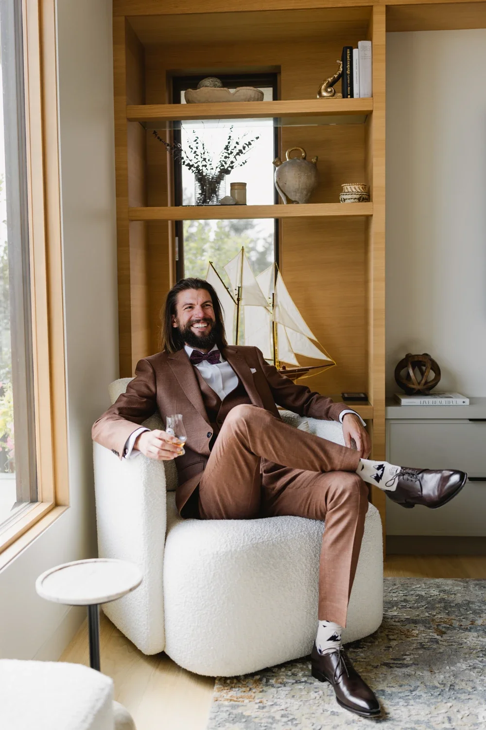 A man in a brown suit sitting in a cream-colored armchair, smiling and holding a glass of whiskey in a cozy room with wooden shelves and nautical decor.