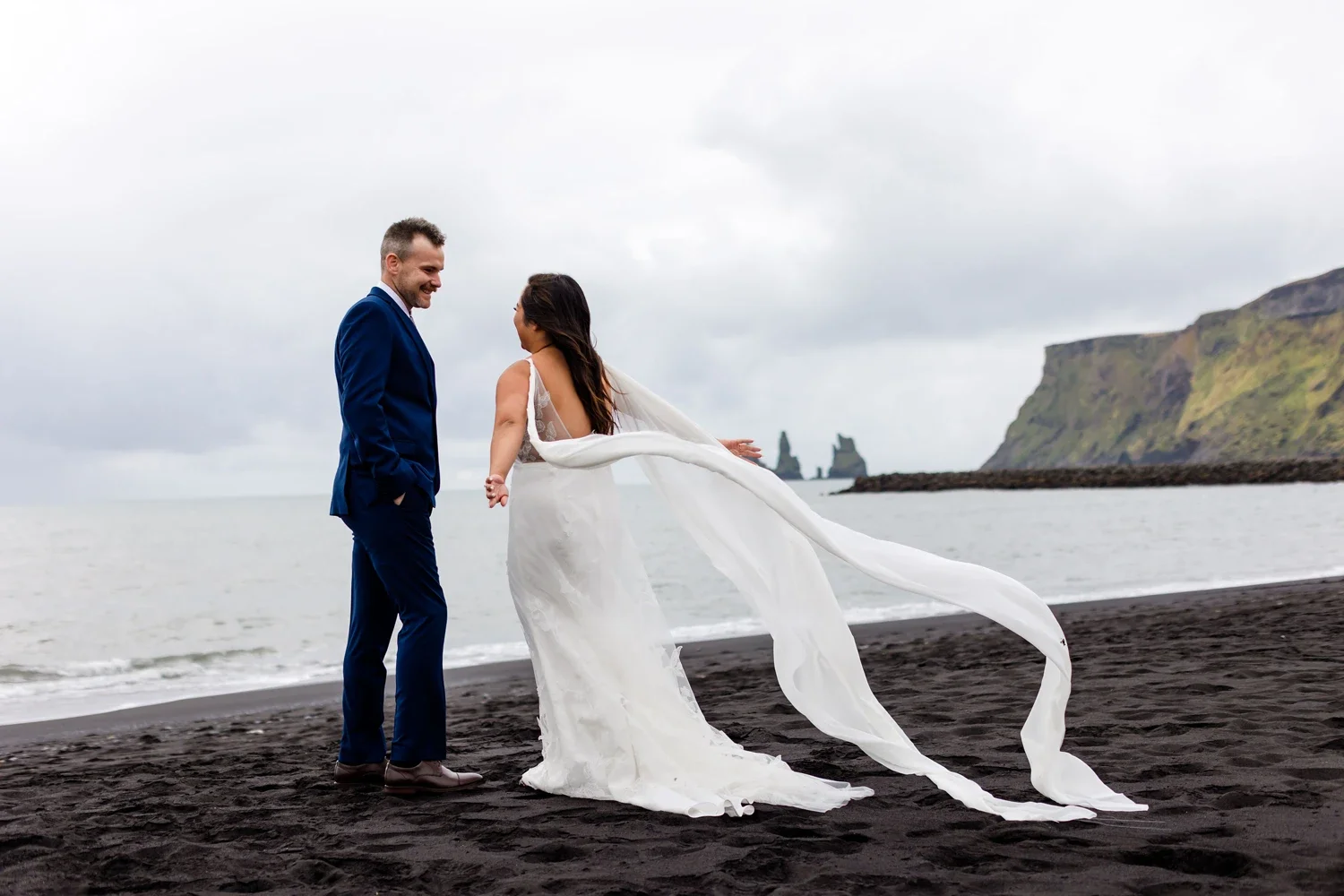 A newlywed couple standing on a black sand beach, holding hands and smiling at each other with cliffs and sea stacks in the background under cloudy skies.