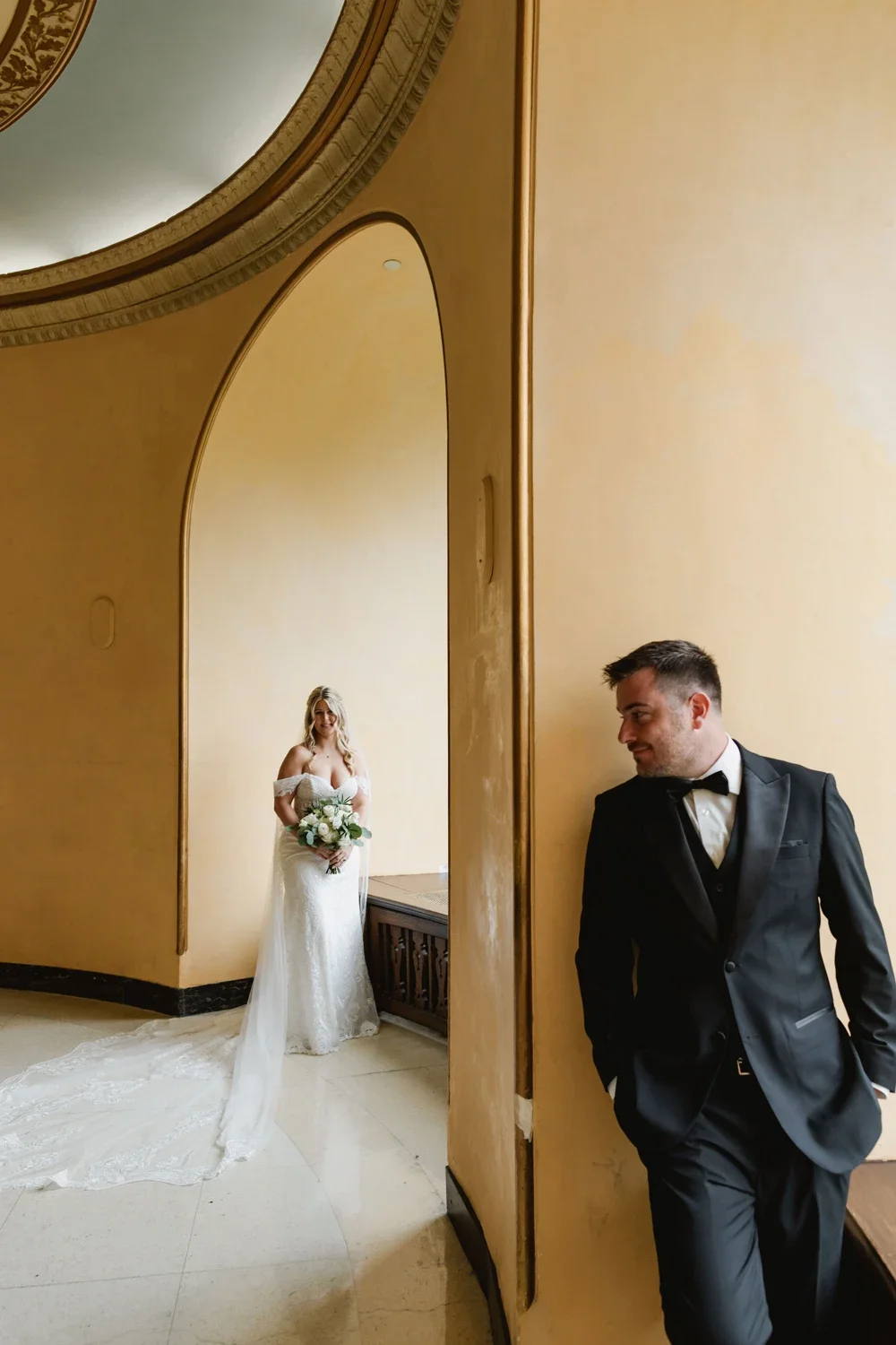 A bride in a white wedding dress holding a bouquet of flowers standing in front of a yellow wall, with a groom in a black tuxedo leaning against a wall, looking at her. The photo is taken through an arched mirror or window frame.