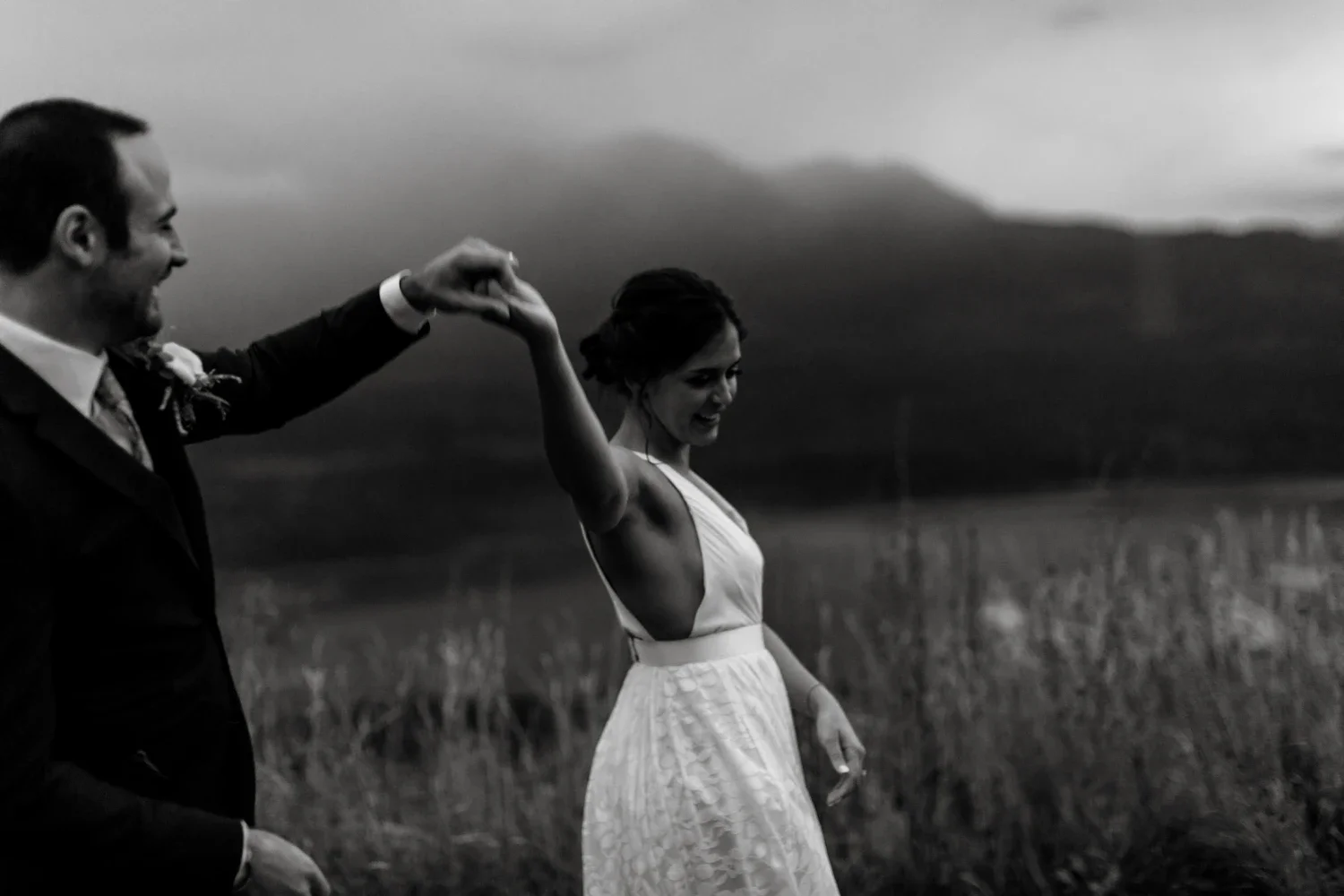 A black and white photo of a man and woman dancing outdoors, with mountains and cloudy sky in the background. The man is wearing a suit, and the woman is wearing a dress.