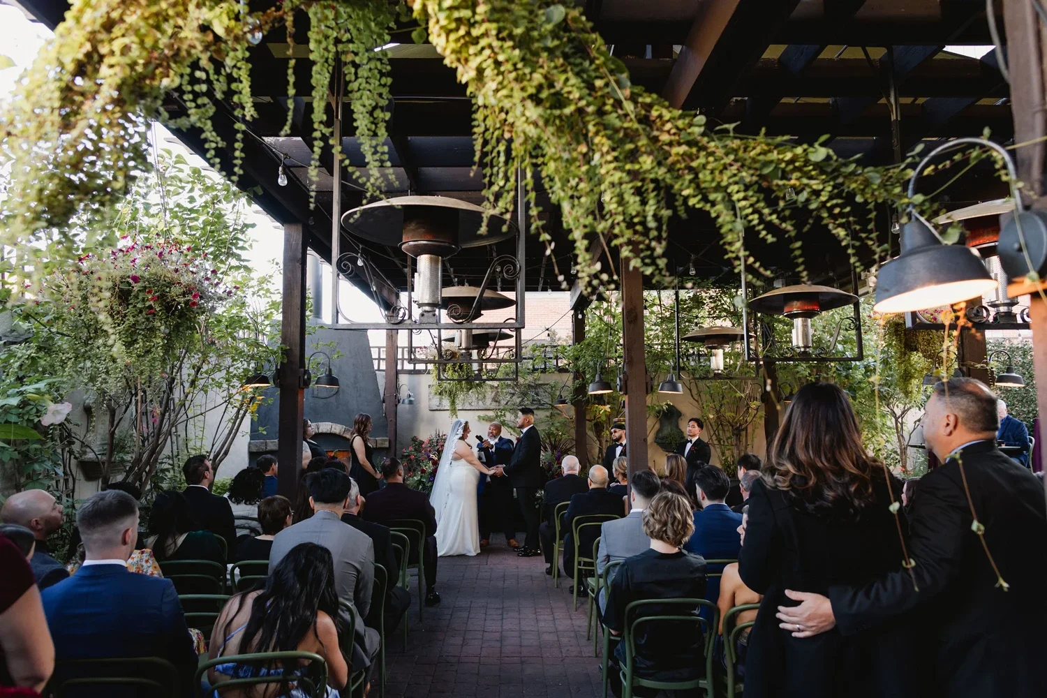 Wedding ceremony taking place outdoors under a canopy with greenery and hanging plants; bride and groom exchanging vows in front of guests seated in rows.