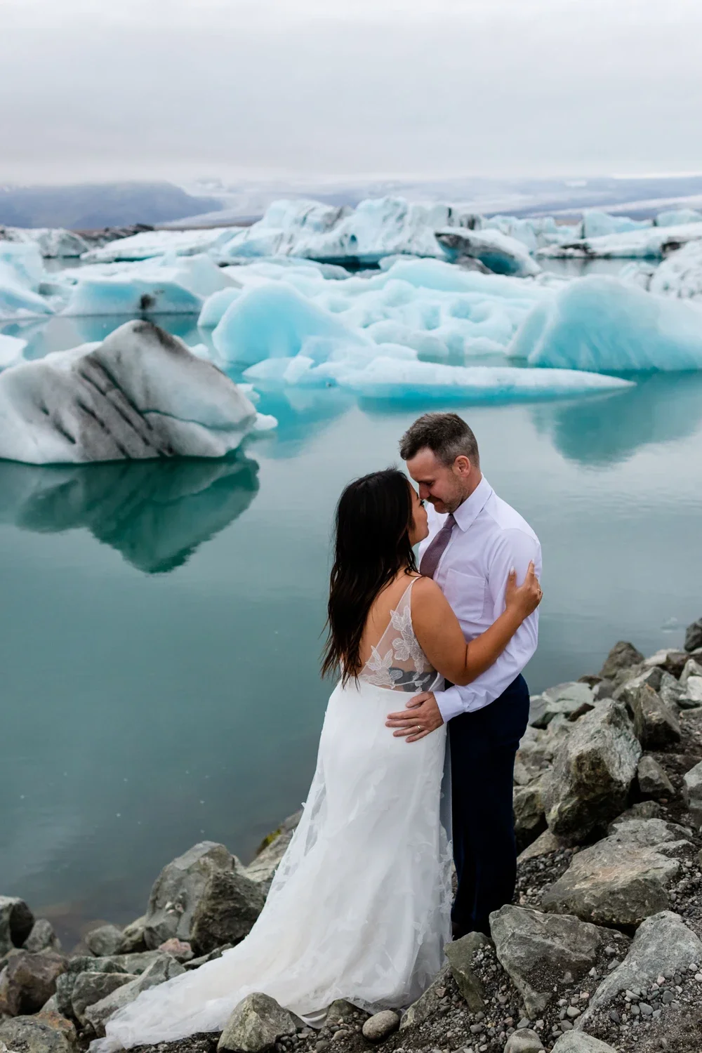 A couple in wedding attire embracing on rocky shores with a glacier and icy landscape in the background.