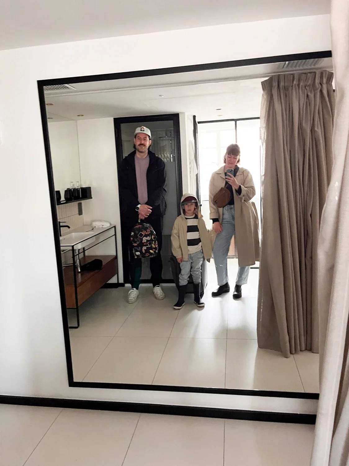 A family of three standing in front of a mirror, taking a selfie in a room with white walls and beige curtains, with a kid holding a backpack and wearing a hat.