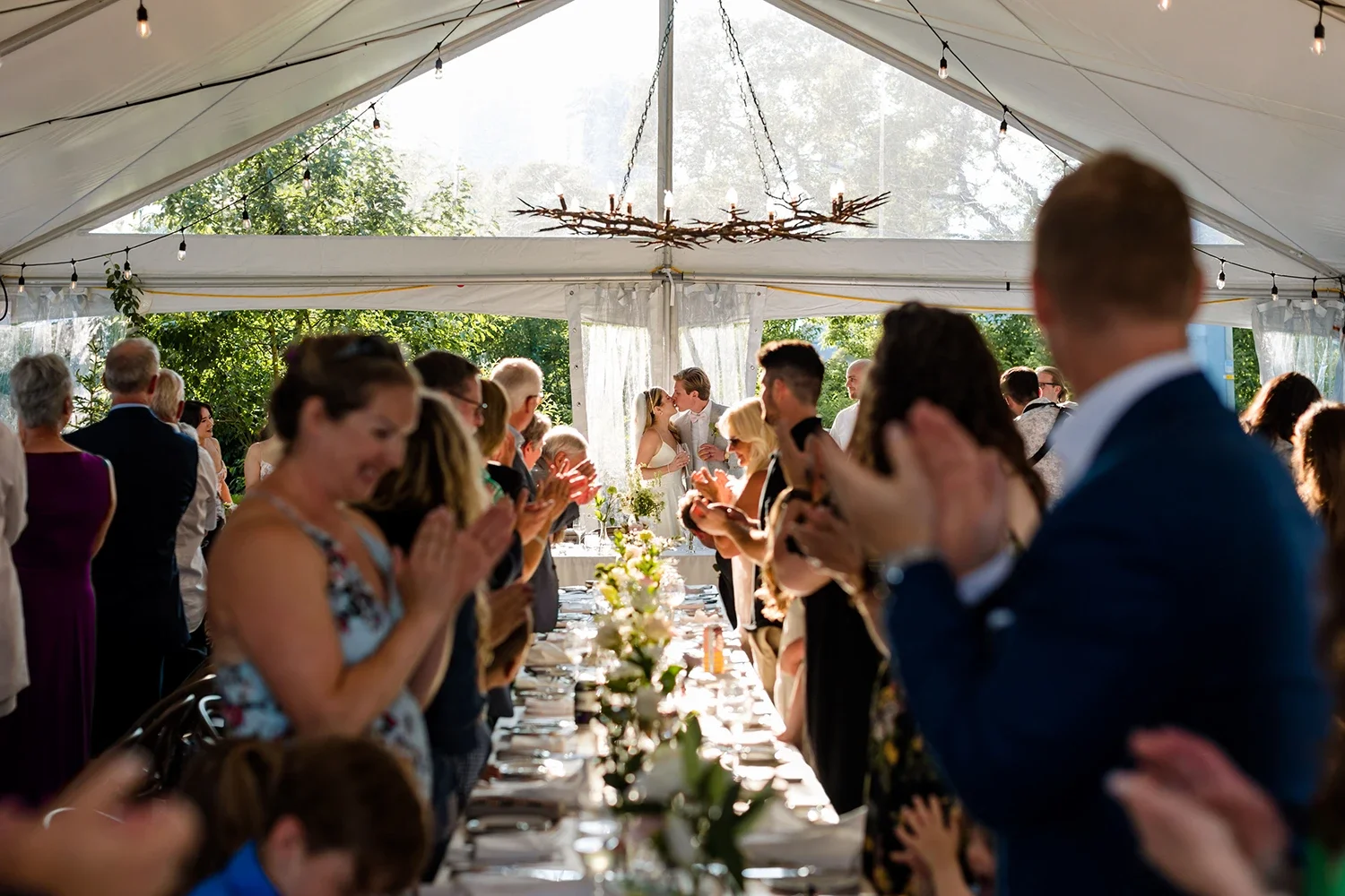 Wedding ceremony inside a celebration tent with guests standing and clapping while the bride and groom kiss at the altar, decorated with a rustic chandelier and string lights.