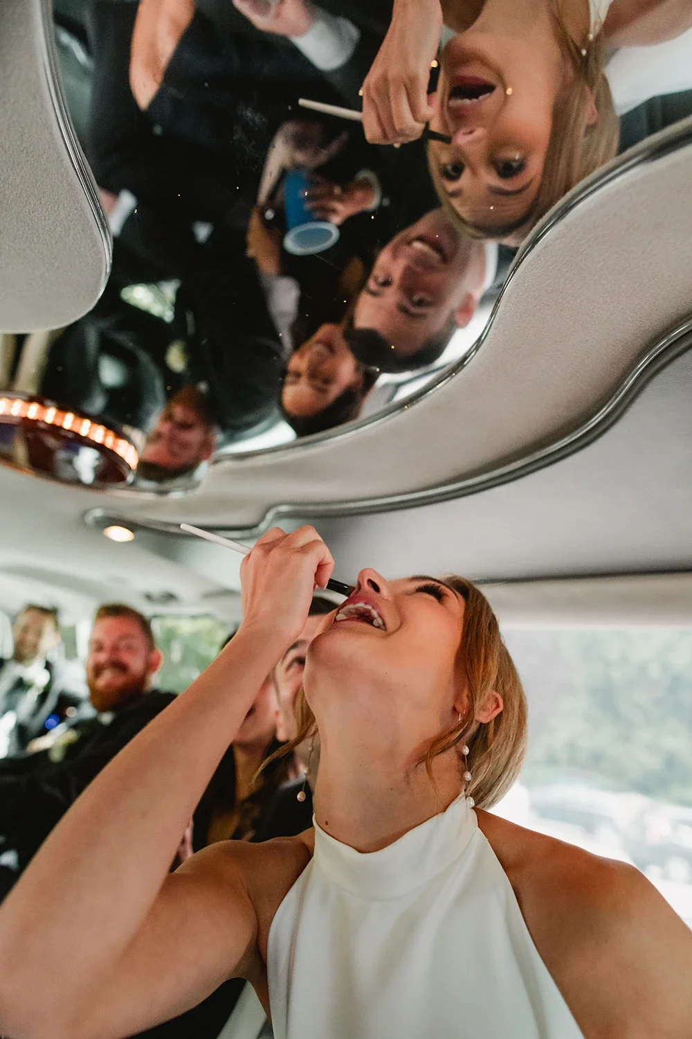 A woman in a white dress is applying makeup with a brush on her chin inside a vehicle, with a group of people, including a man with a beard, visible in the background.