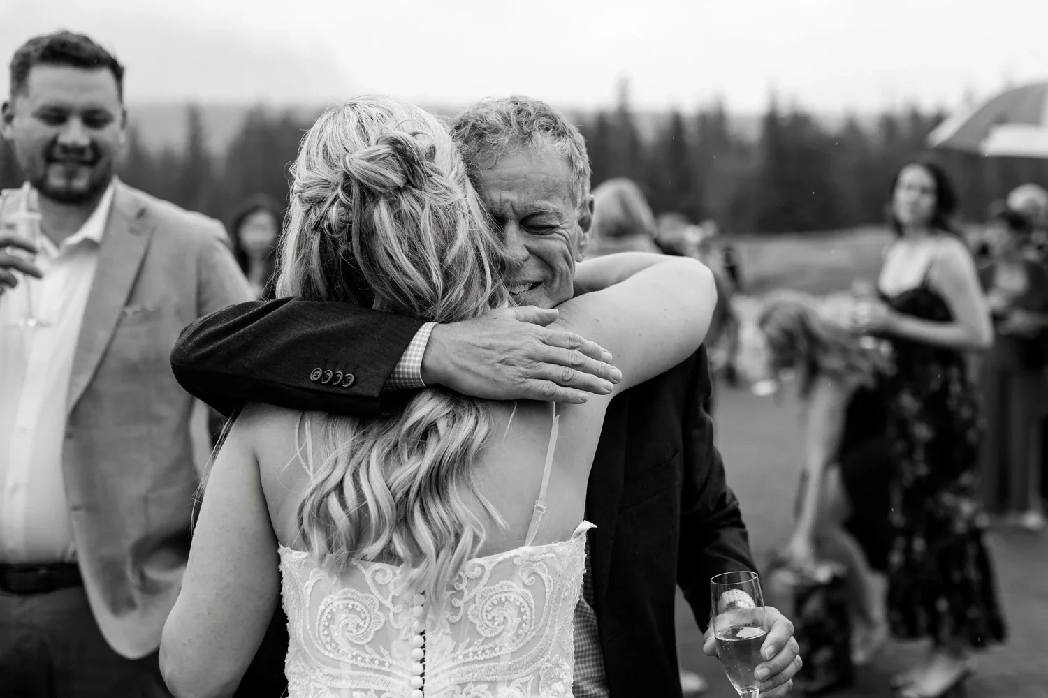 A woman and an elderly man hug tightly at an outdoor celebration, with a woman holding a glass of champagne in the background.