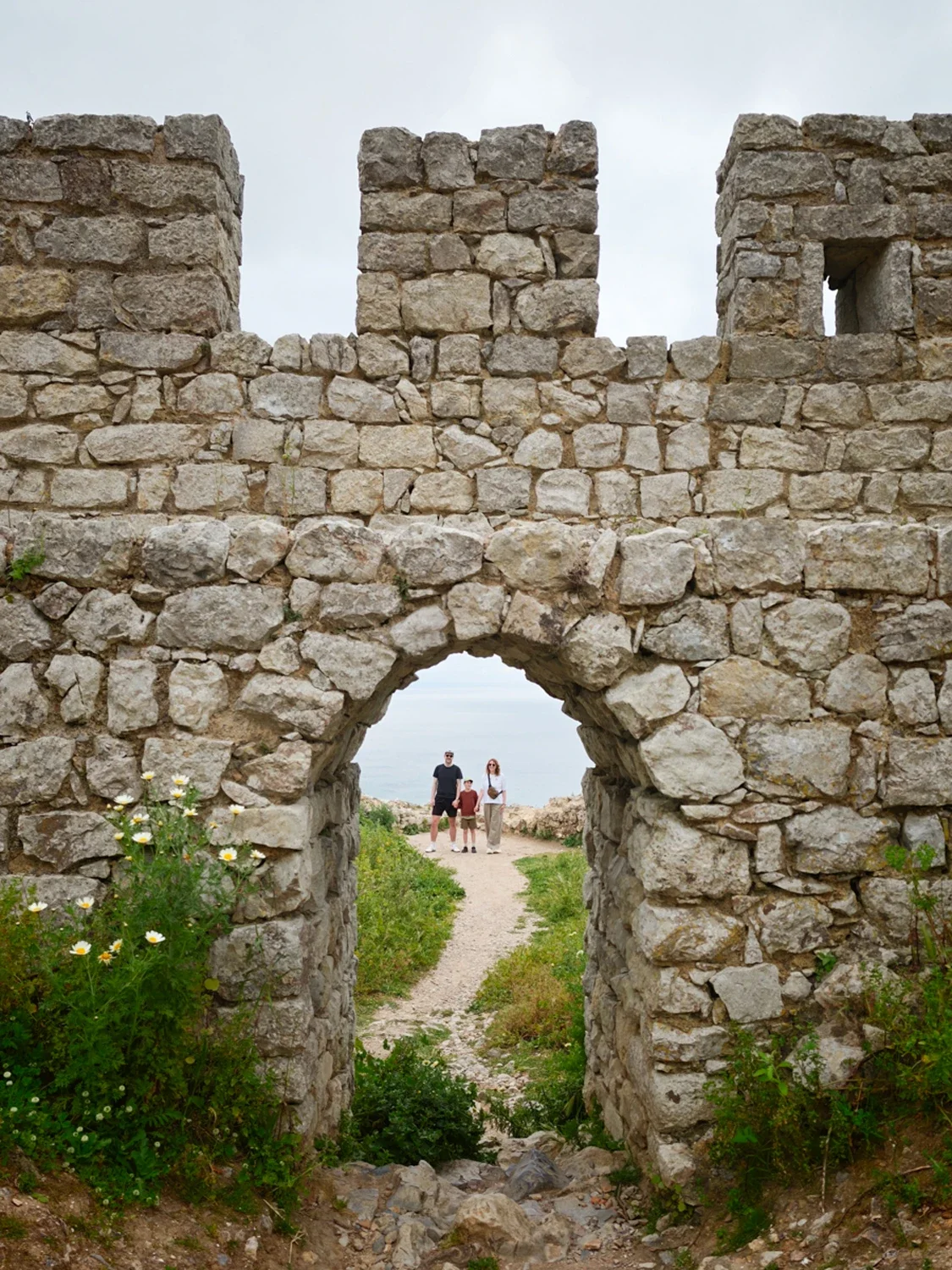 Ancient stone fortress with a small arched gateway, three people walking through it, greenery around the base, and an open field in the background.