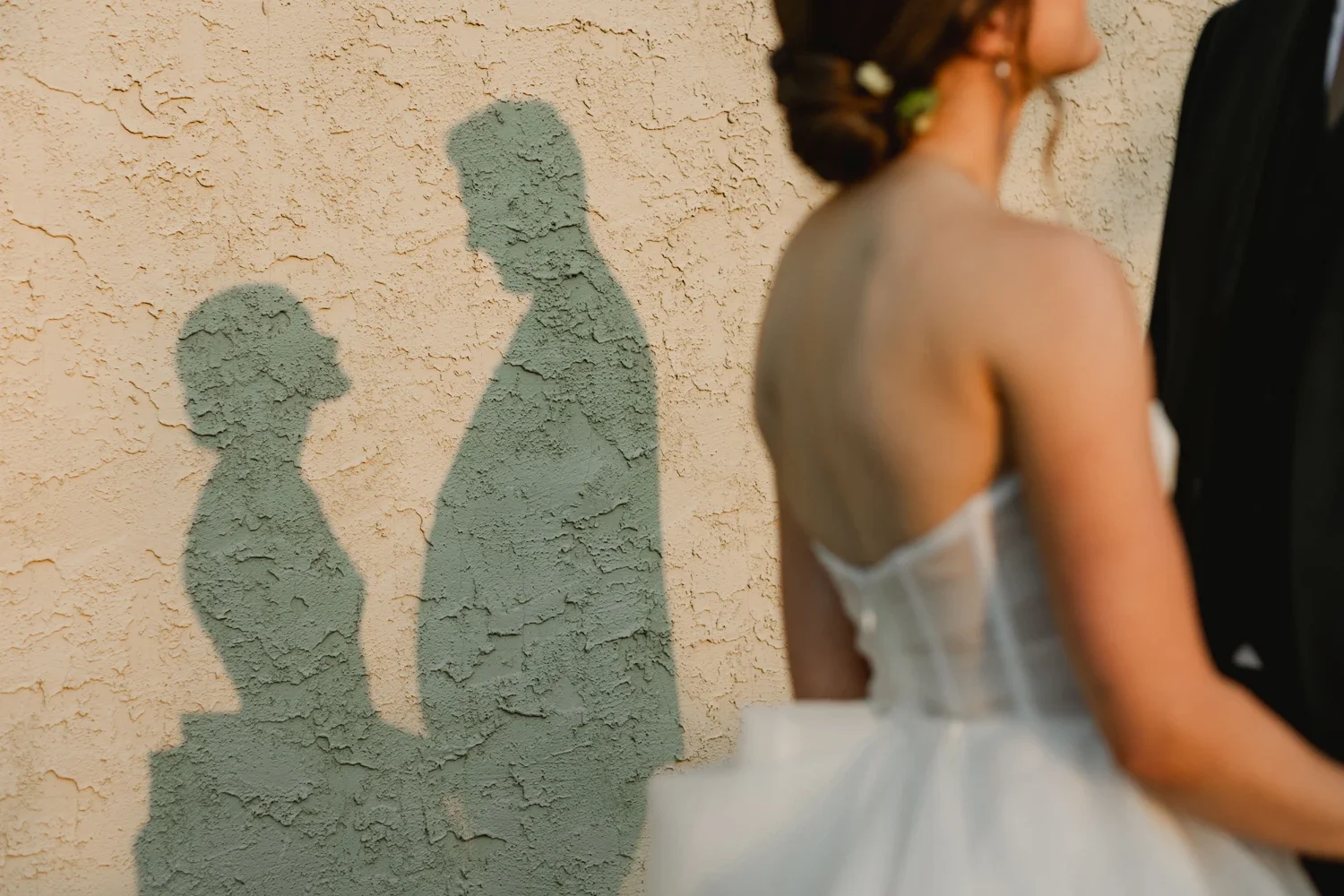 Shadow of a woman in a wedding dress and a man in a suit on a textured beige wall, with partial view of the bride