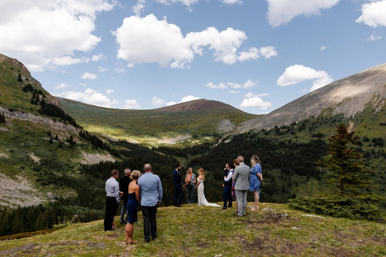 A wedding ceremony taking place outdoors on a grassy hilltop surrounded by mountains and forest under a partly cloudy sky.