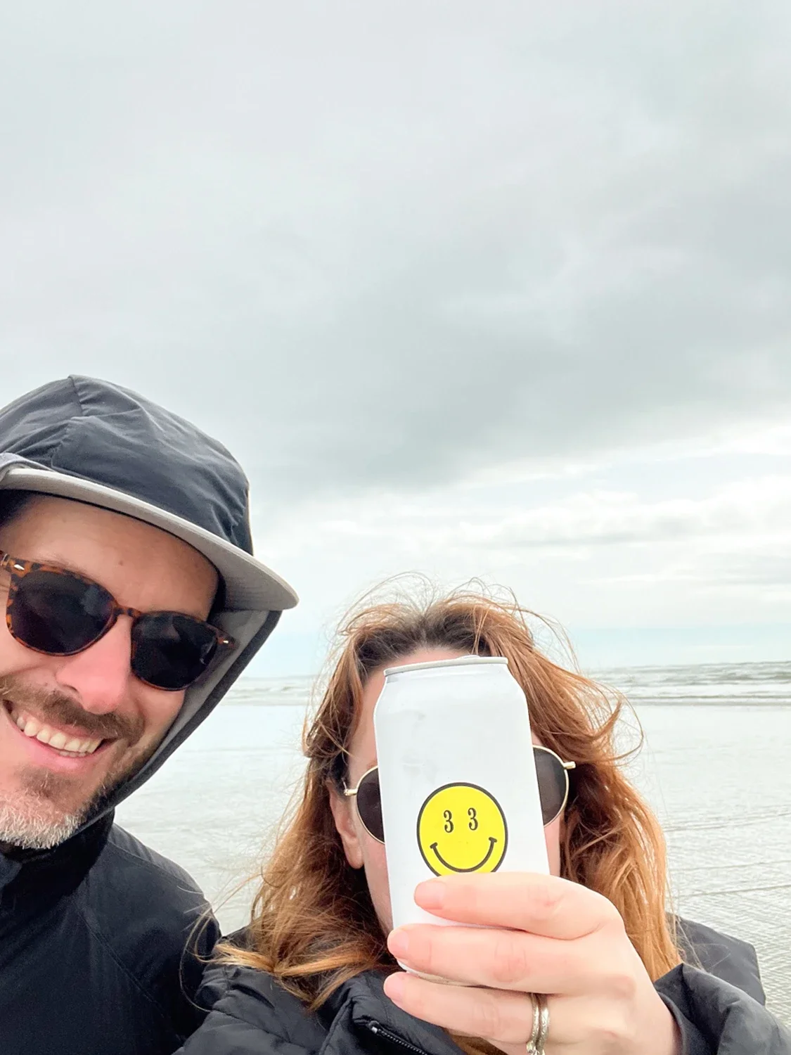 A smiling man and woman taking a selfie on the beach. The woman is holding a white can with a yellow smiley face sticker over her face, and both are wearing sunglasses and jackets. The sky is cloudy and the ocean is in the background.