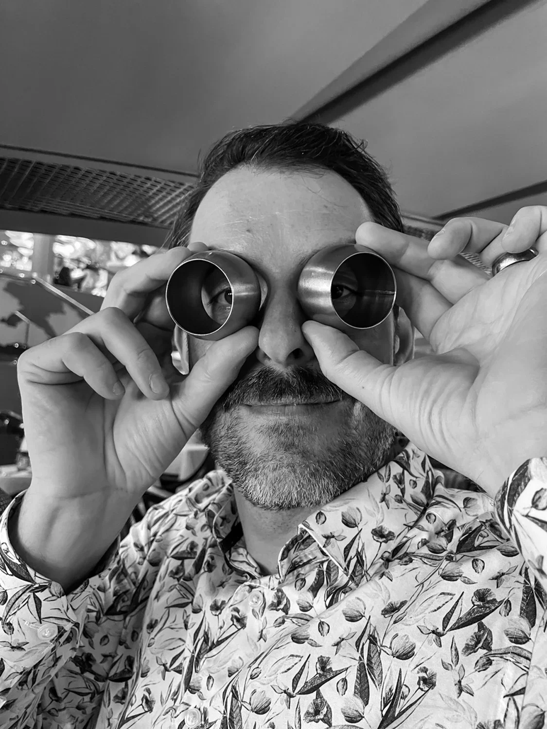 A man with a beard and floral shirt looking through metal optical tubes, holding them to his eyes, in black and white.