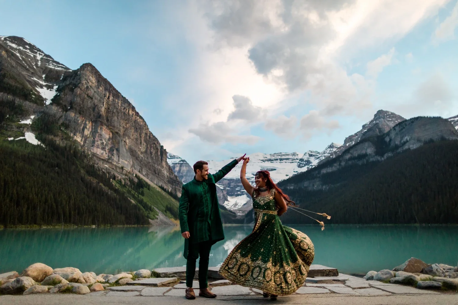 A man and woman dancing together by a mountain lake, with snow-capped peaks and a cloudy sky in the background. The woman is wearing a green traditional dress, and the man is dressed in dark green attire.