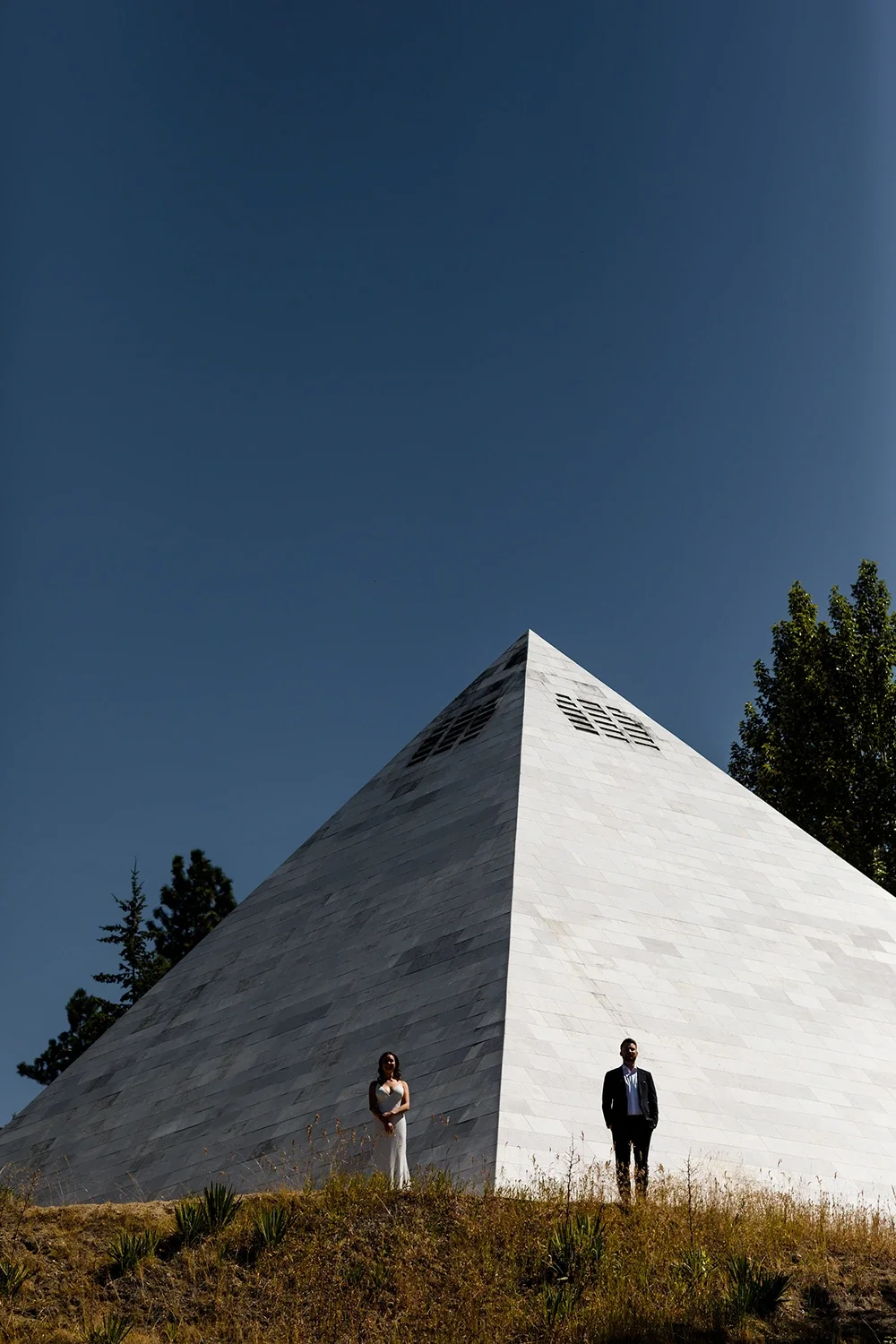 A modern, angular white building with small windows is seen from below against a clear blue sky. Two people, a woman in a white dress and a man in a dark suit, stand on dry grass at the base of the building, with trees in the background.