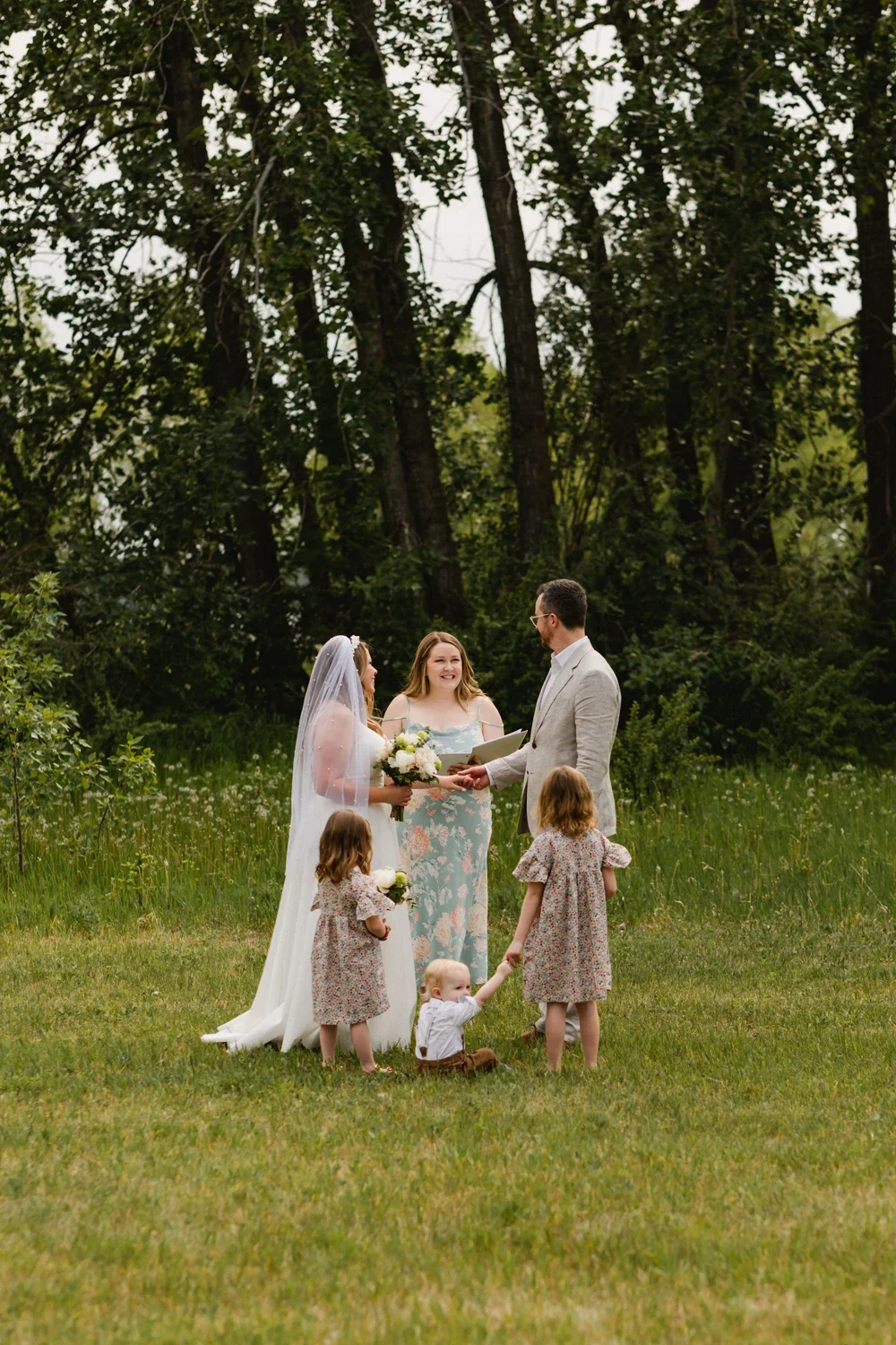 A wedding ceremony outdoors with a bride, groom, officiant, and three children, set in a green, wooded area.