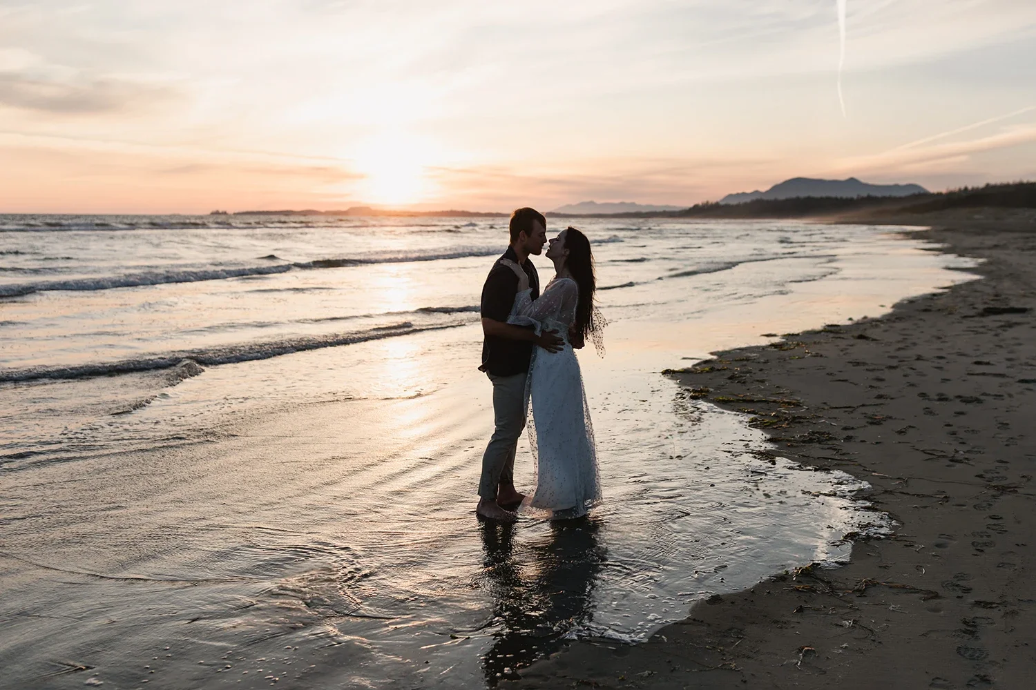 A couple is standing close together at the beach during sunset, holding each other and looking into each other's eyes, with the ocean and mountains in the background.
