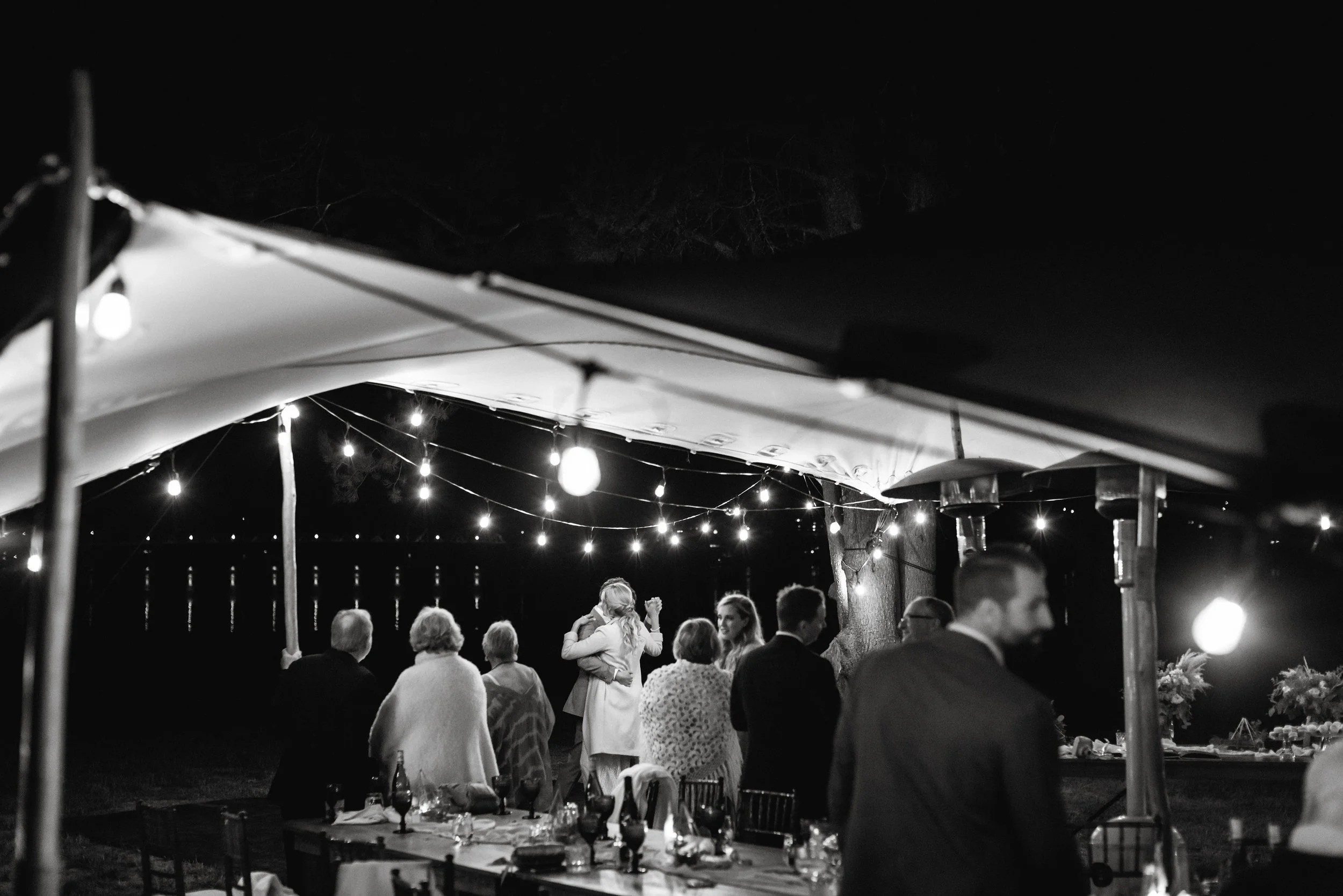 Nighttime outdoor gathering with string lights, guests, and tables, under a canopy.
