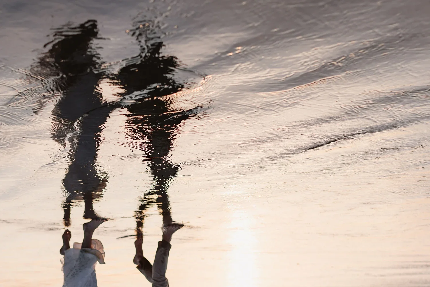 Two children holding hands and walking on a beach with wet sand reflecting the sunset sky.