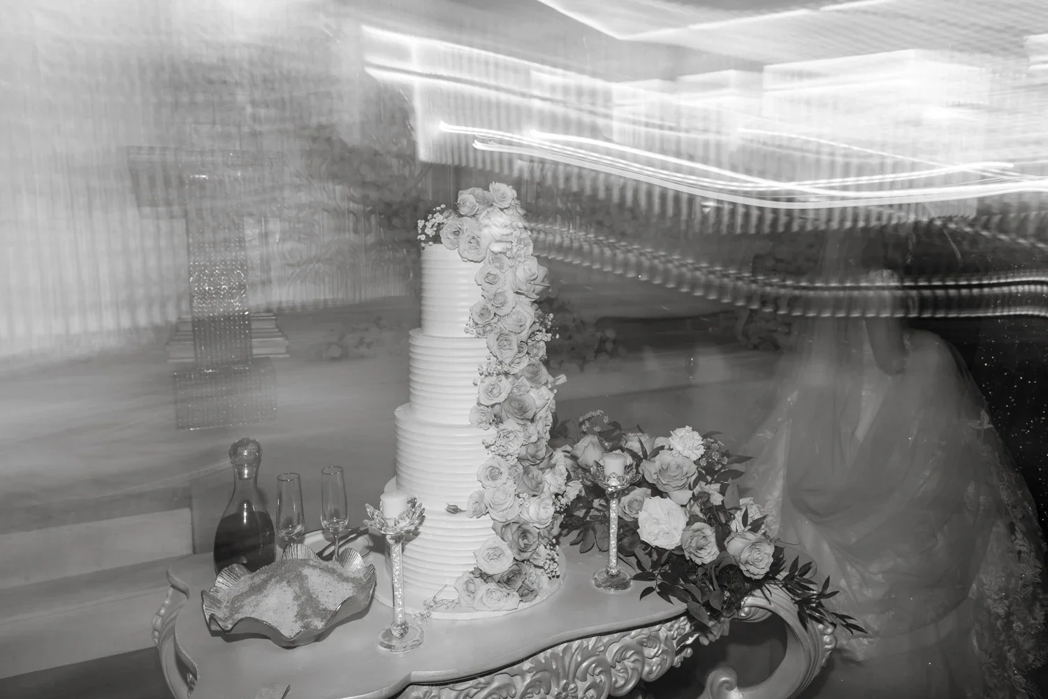 A wedding cake decorated with flowers, placed on an ornate table with wine glasses, a bottle, candles, and a decorative tray, at a wedding reception.