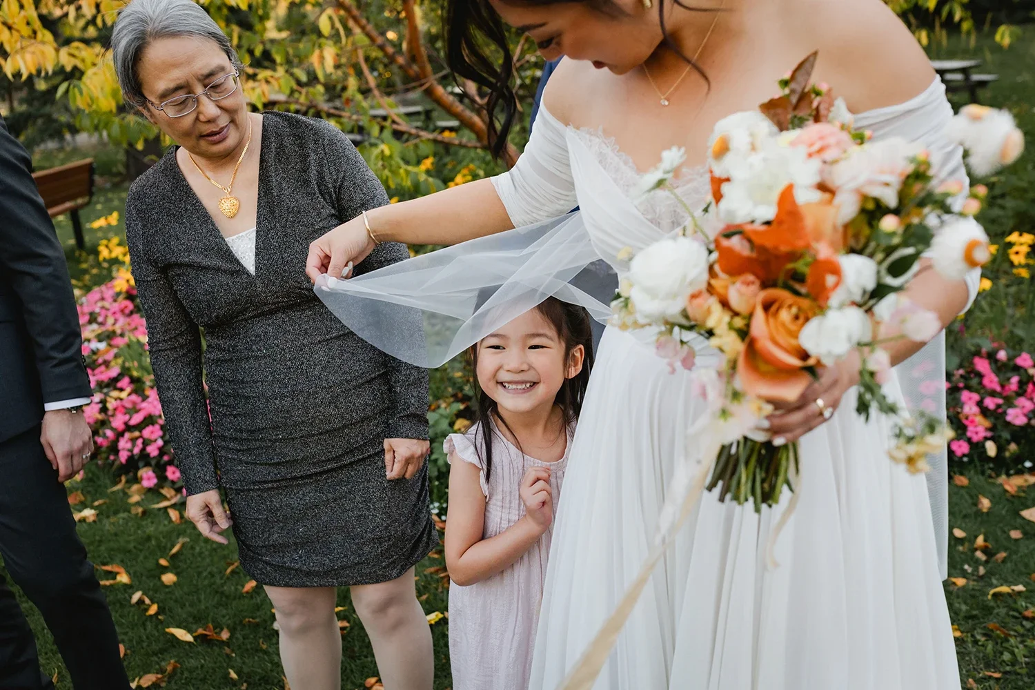 A woman in a wedding dress holding a bouquet of flowers is smiling and looking down at a young girl smiling underneath her veil at an outdoor wedding.