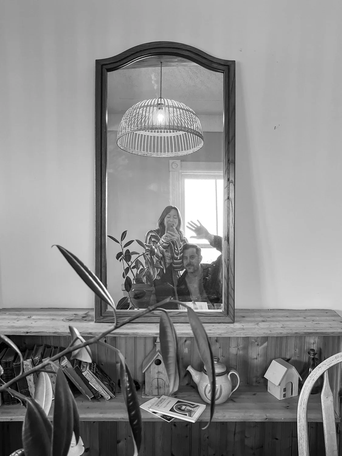 A mirror hanging above a wooden shelf reflects a woman and a man taking a selfie, with the woman smiling and the man waving. A plant, a teapot, books, a picture, and a small house model are on the shelf, and a window is visible behind them.