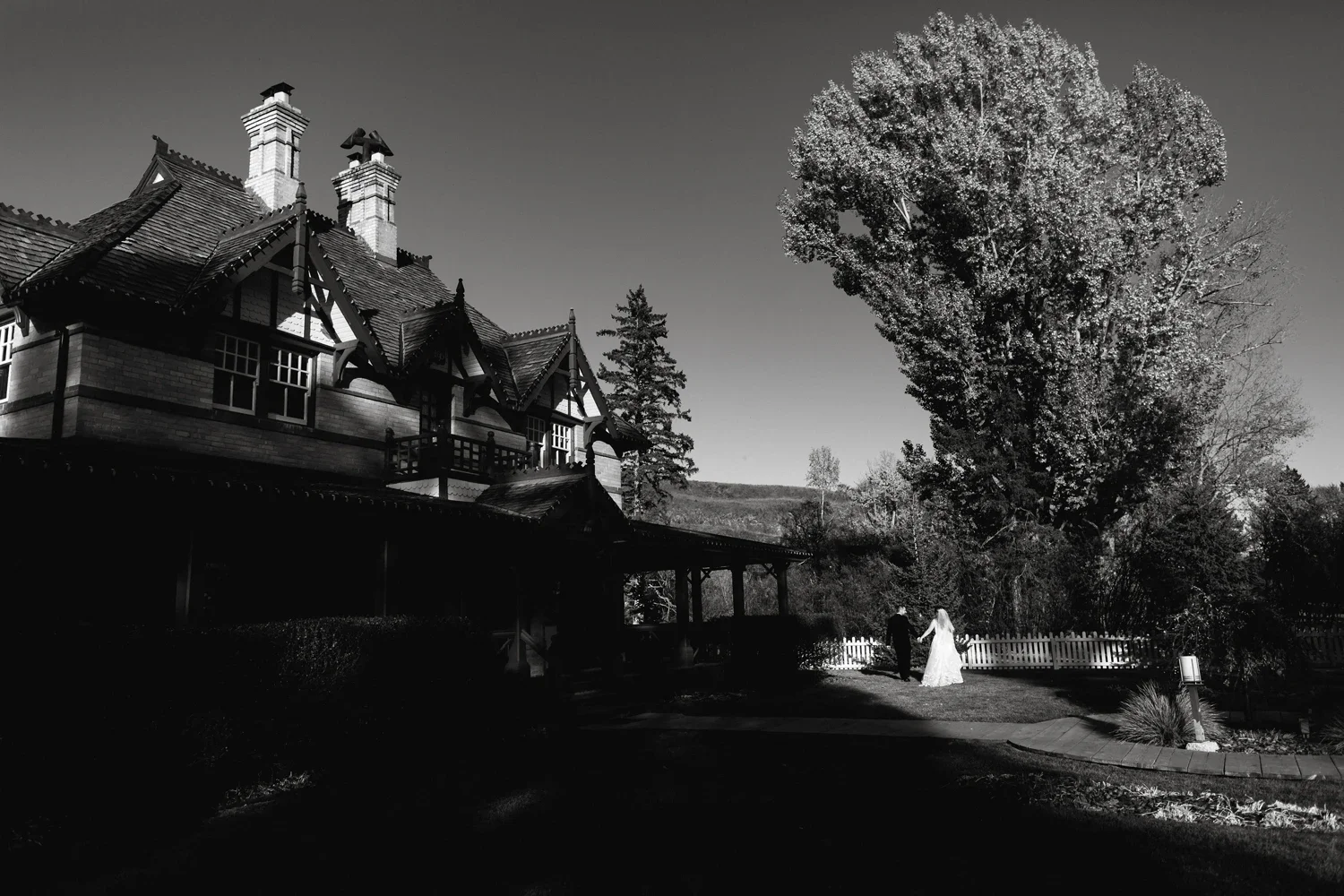 A black and white photo of a large, historic house with multiple chimneys and ornate roofing, surrounded by tall trees. In the foreground, a couple dressed in wedding attire is walking hand in hand near a white picket fence.