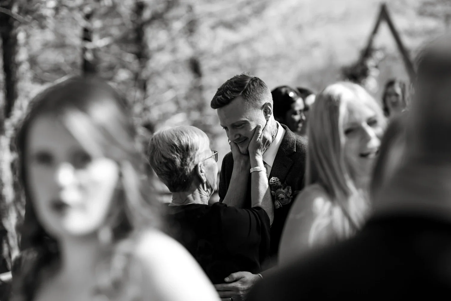 A young man, dressed in a suit, holding an elderly woman's face, both smiling warmly, surrounded by blurred people at an outdoor event.