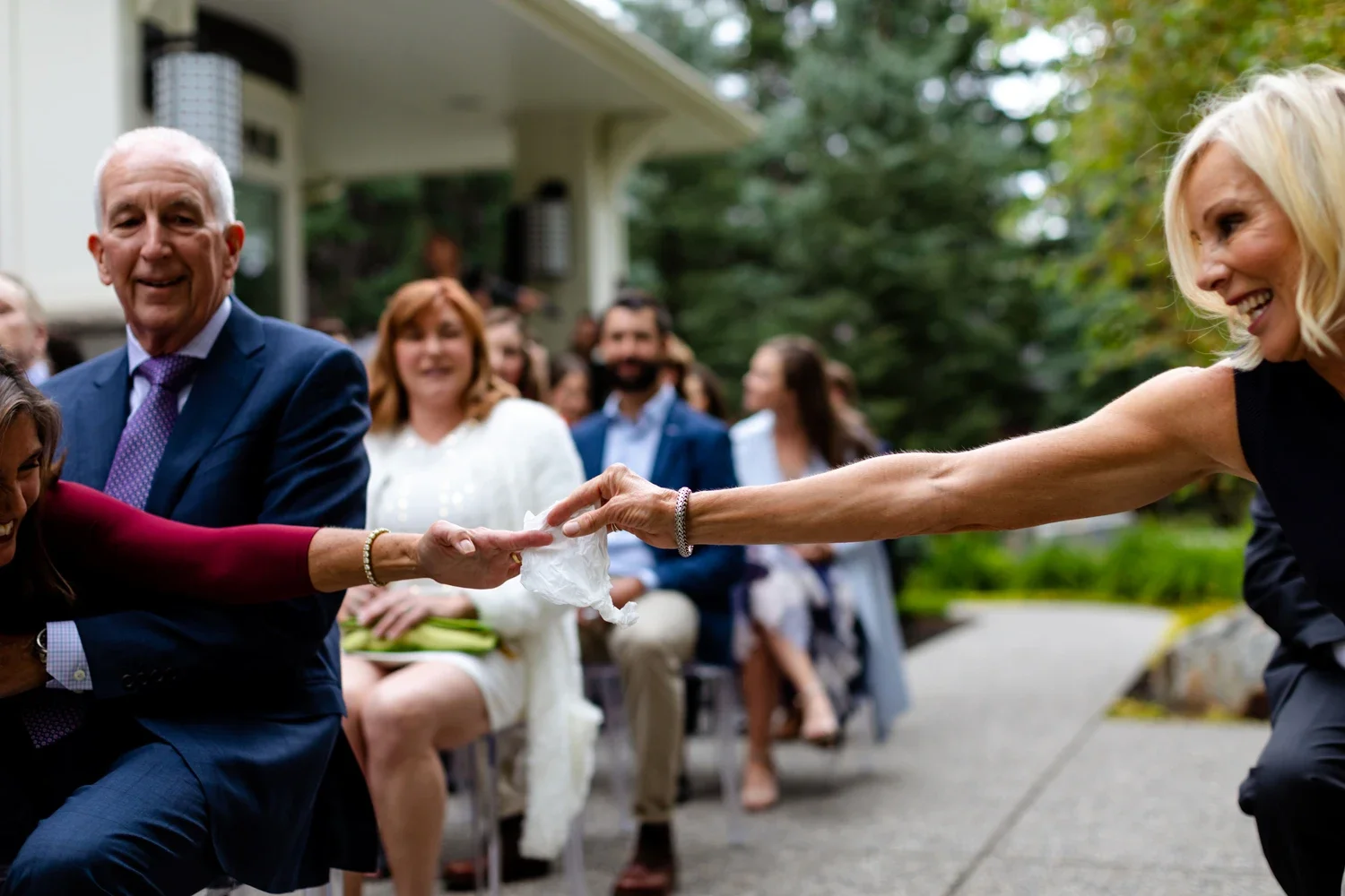 Two women are in a playful tug of war with a tissue while people watch and smile at an outdoor gathering.