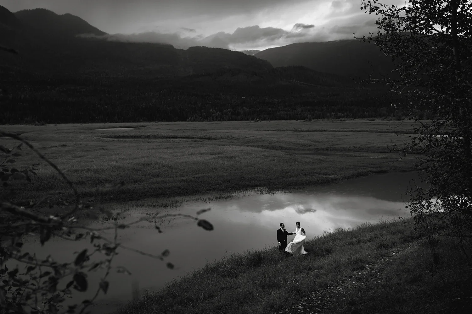 A black and white photograph of a couple in wedding attire standing near a body of water, surrounded by grassy landscape and mountains in the background.