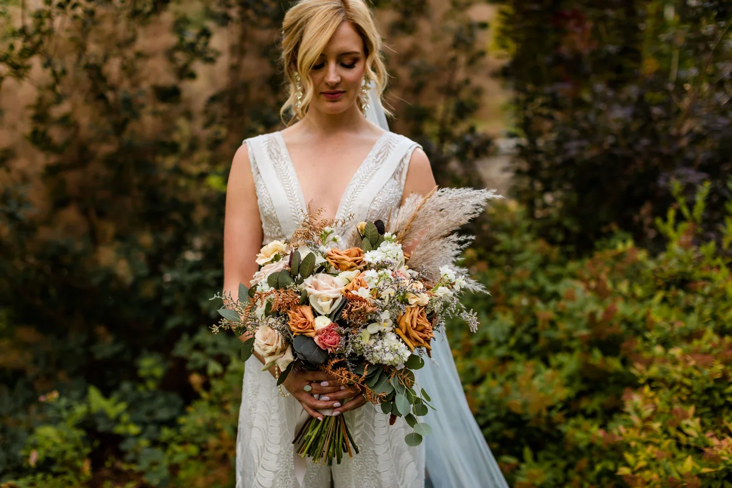 A woman in a white dress holding a bouquet of mixed flowers and dried plants, standing outdoors with greenery and bushes in the background.