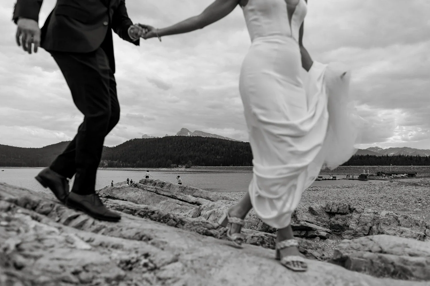 A black and white photograph of a couple holding hands on a rocky beach. The man is dressed in a suit and the woman is wearing a flowing white dress and sandals. There are mountains, water, and a cloudy sky in the background.