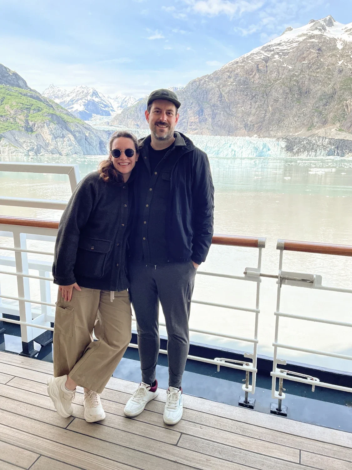 A smiling woman and man standing side by side on a boat with railing, with snowy mountains, a glacier, and water in the background. They are dressed in casual outdoor clothing.