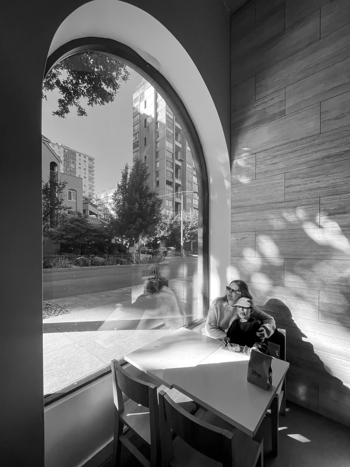 A woman and a young girl sitting at a table inside a cafe, with large arched window showing a city street scene outside, including trees, buildings, and parked cars.