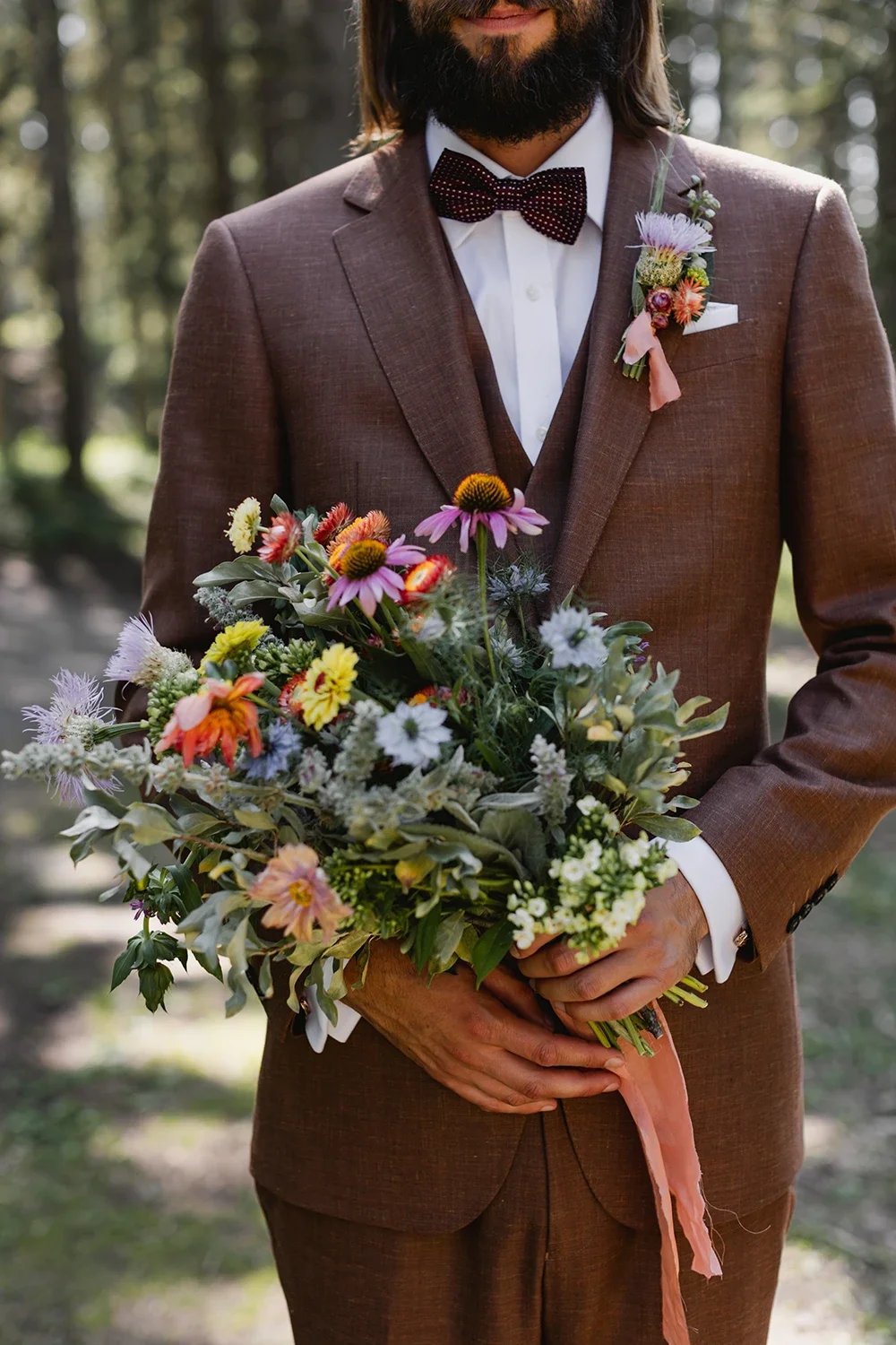 Man in brown suit holding a bouquet of colorful wildflowers, wearing a white shirt, dark polka dot bow tie, and a flower boutonniere, outdoors in a wooded area.