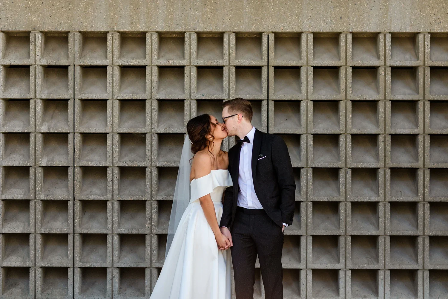 Bride and groom kissing in front of a concrete grid wall, holding hands.
