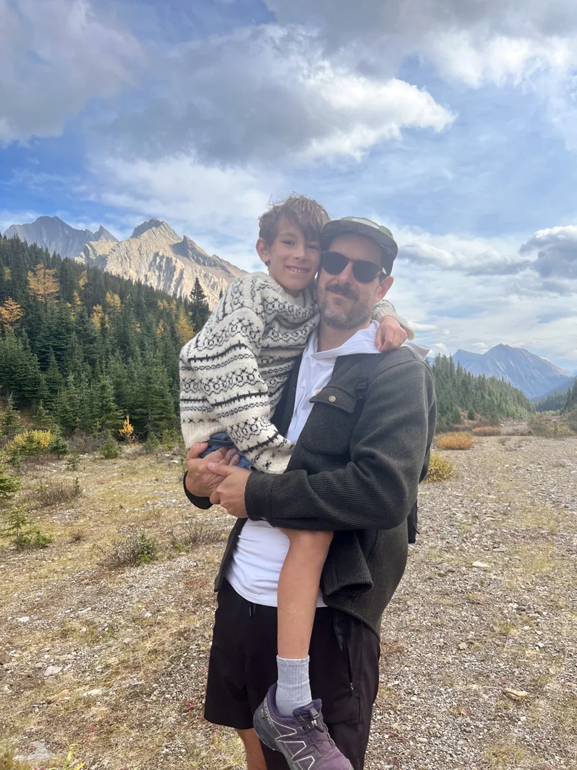 A man in sunglasses and a dark jacket holds a smiling boy in an alpine landscape, with mountains and a cloudy sky in the background.