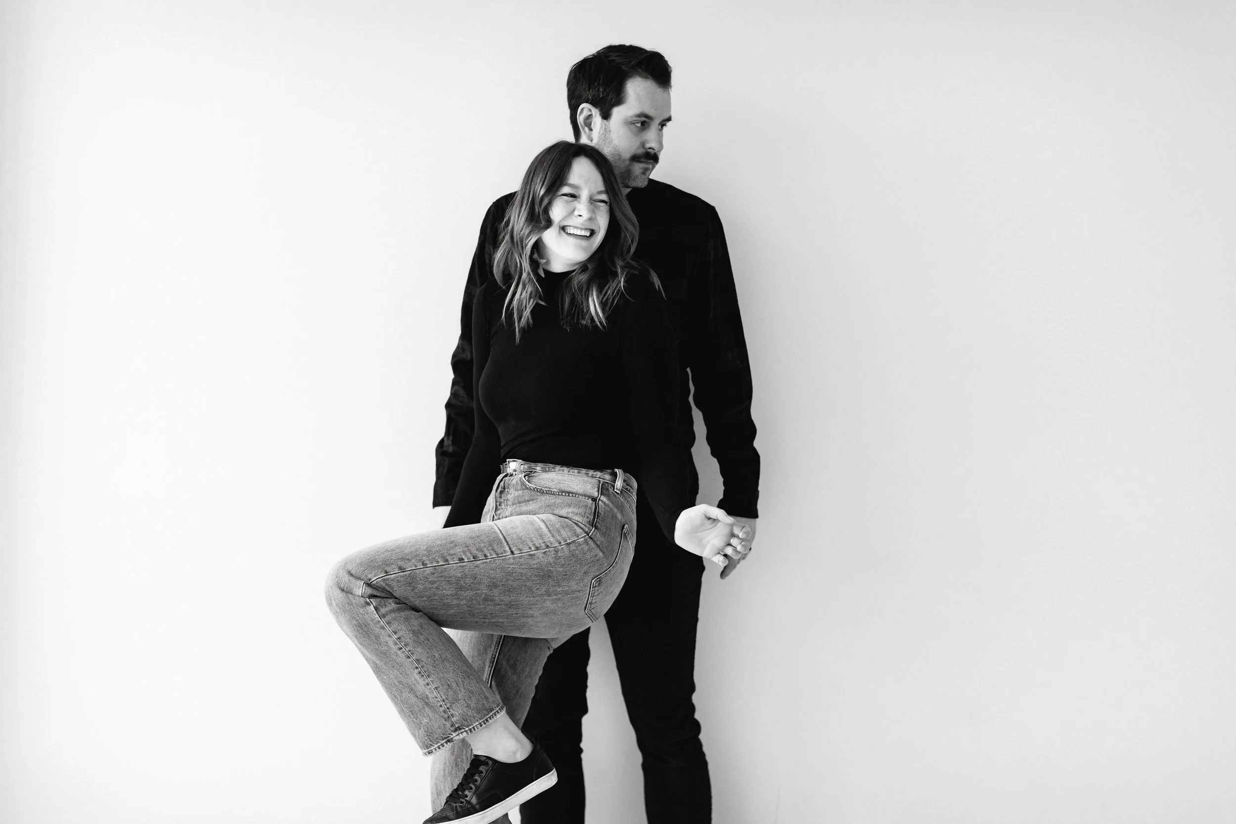 Black and white photo of a man standing behind a woman, who is smiling and laughing with her eyes closed. The woman is wearing a black top and jeans, and the man is wearing a dark shirt. The woman's leg is lifted and bent at the knee as if she is jumping or dancing.
