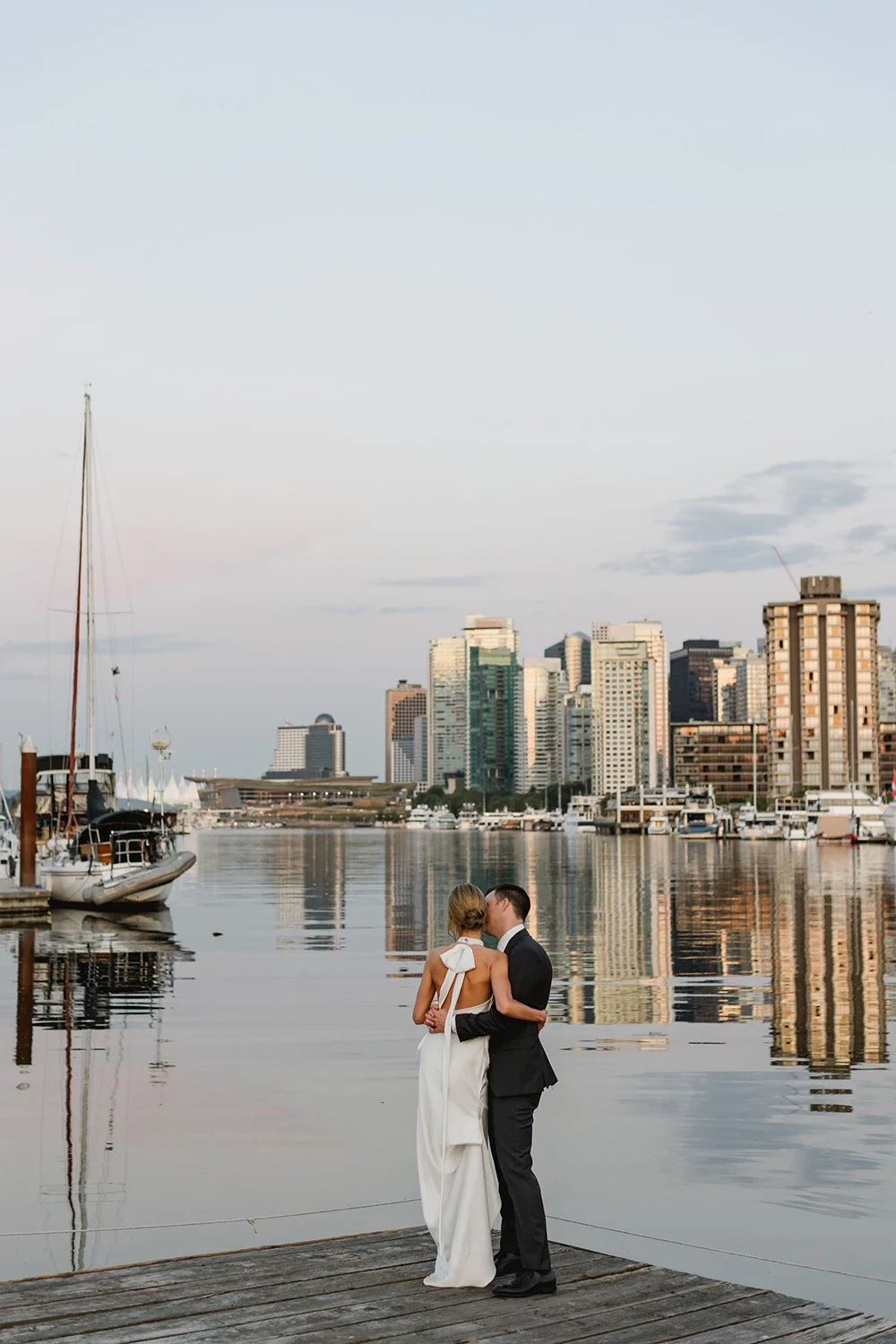 A couple dressed in wedding attire sharing a kiss on a wooden dock by the water with city skyline and boats in the background.