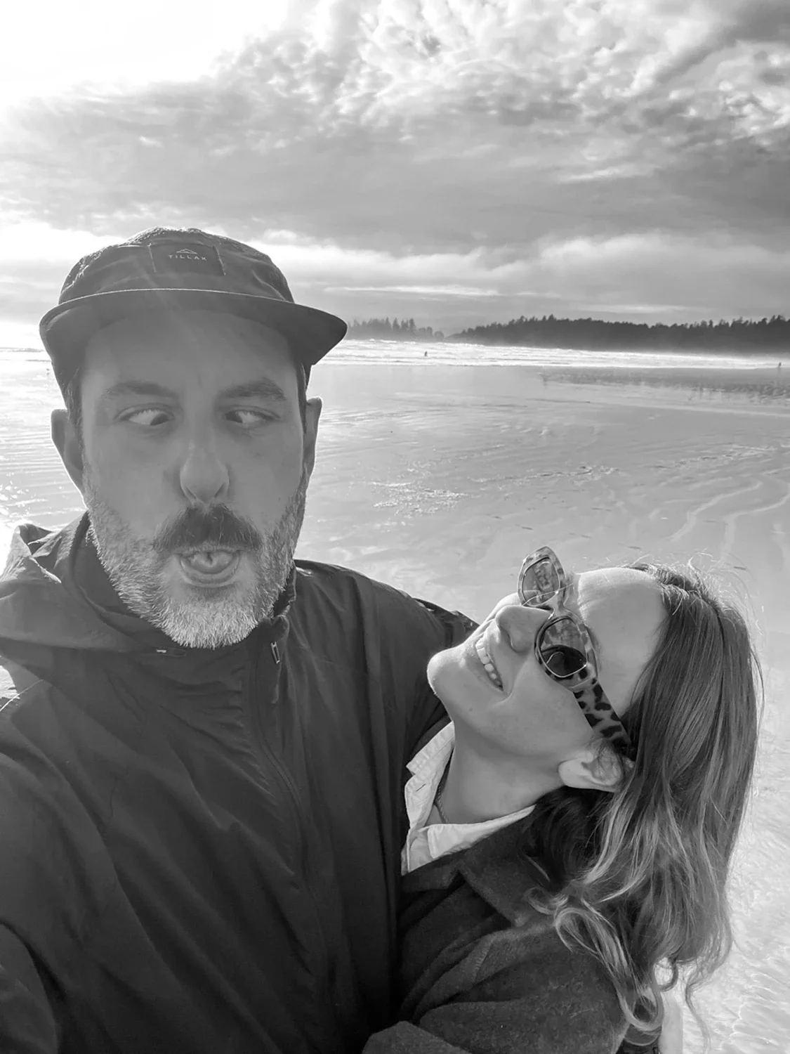 A man and woman taking a selfie on the beach. The man is wearing a cap and a jacket, and the woman is wearing sunglasses and a jacket. They are smiling and appear to be enjoying a sunny day with clouds and trees in the background.
