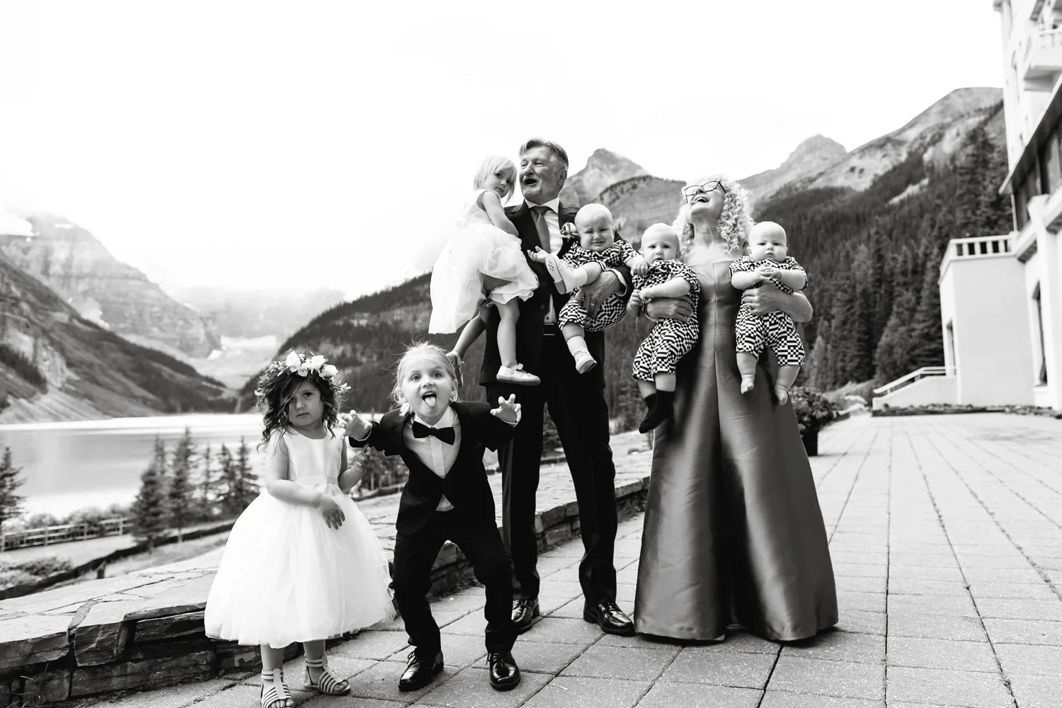 A family dressed in formal attire posing outdoors on a mountain backdrop. The family includes two women, one man, and five children, some in dresses and tuxedos, with mountains and a lake in the background.
